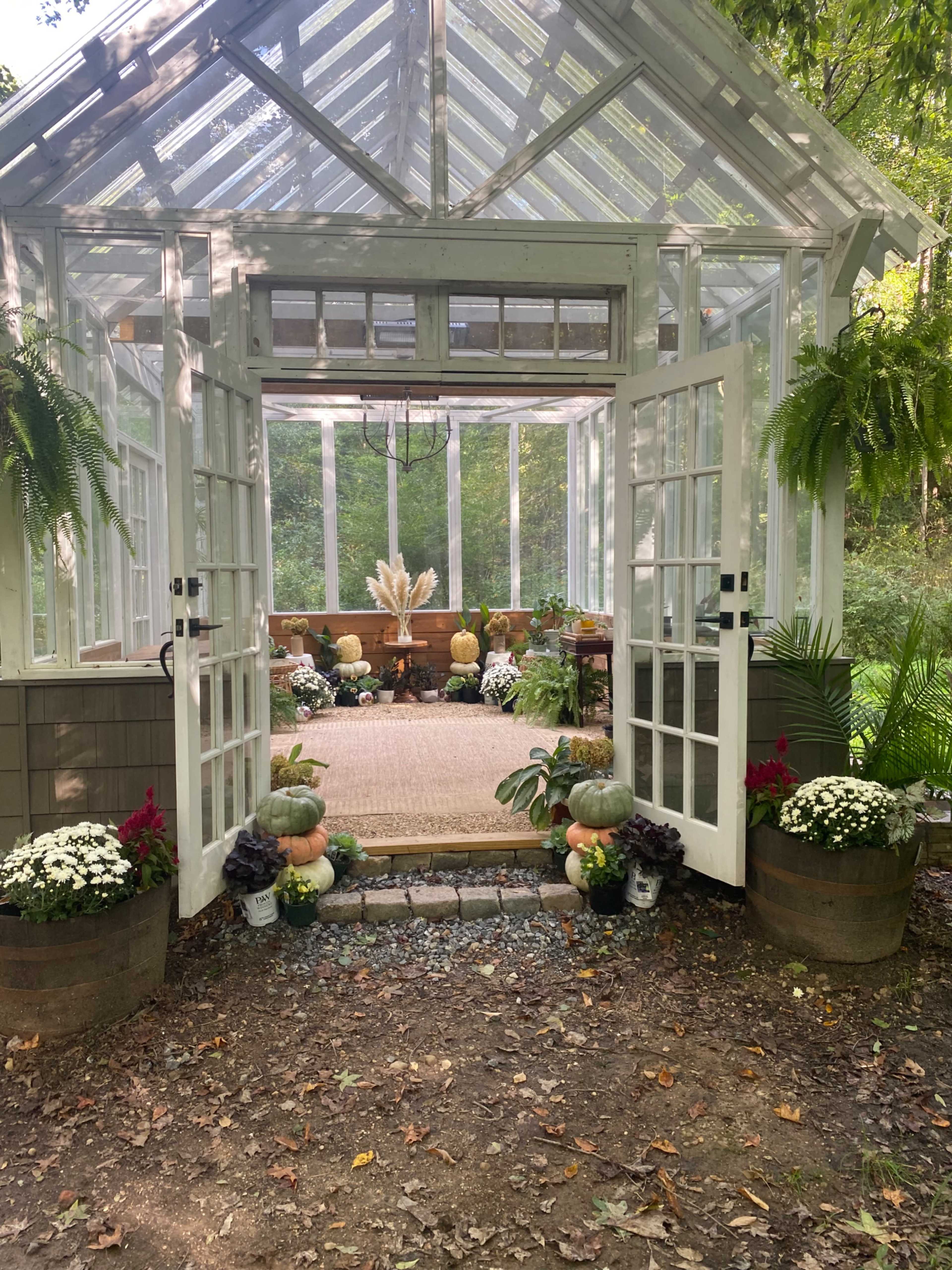 A glass greenhouse with open French doors displays various plants and decorative elements on a stone pathway.