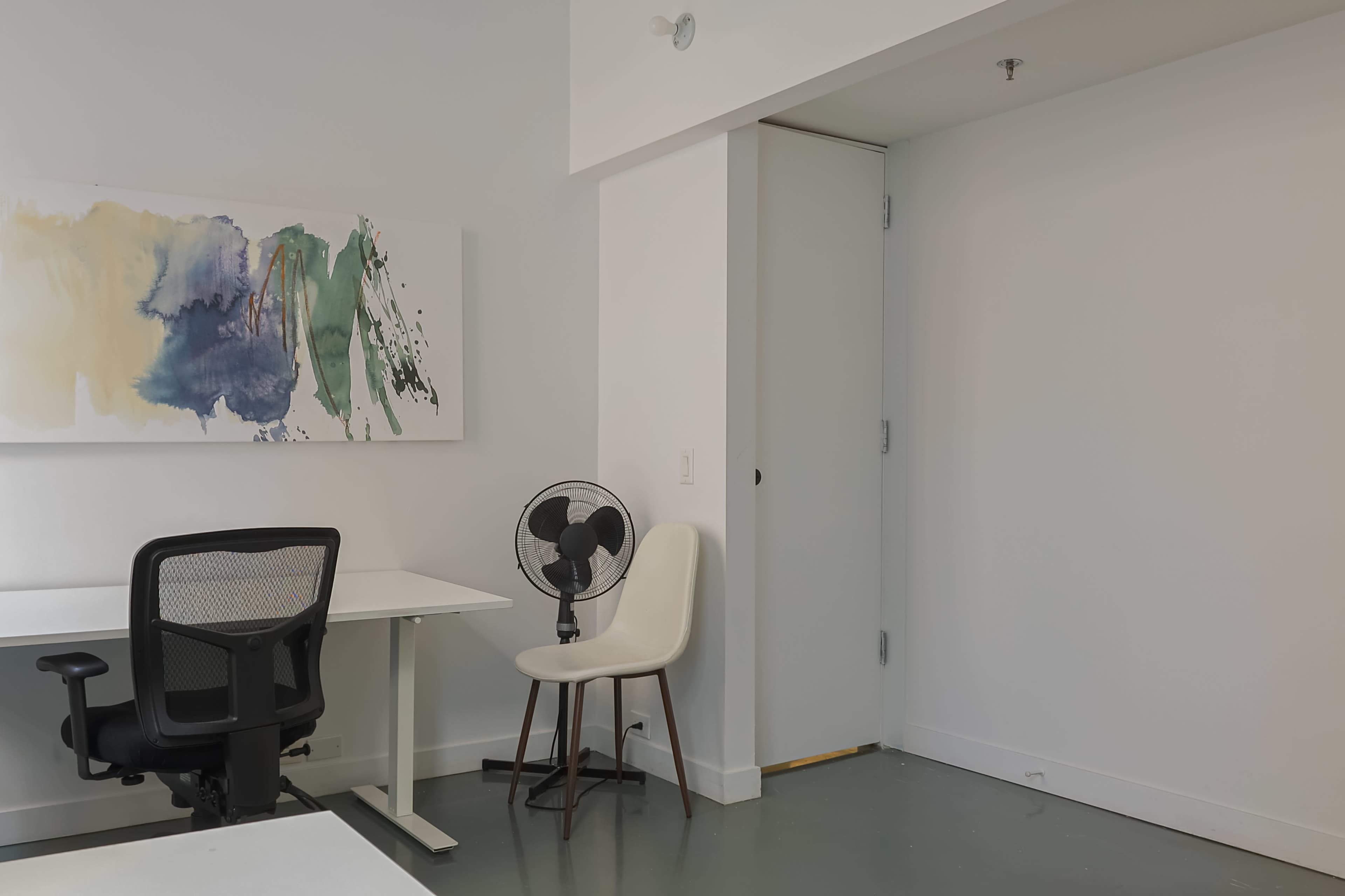 The image shows a minimalist office space with a desk, a black office chair, a white chair, a wall-mounted painting, and a small fan next to a closed door.