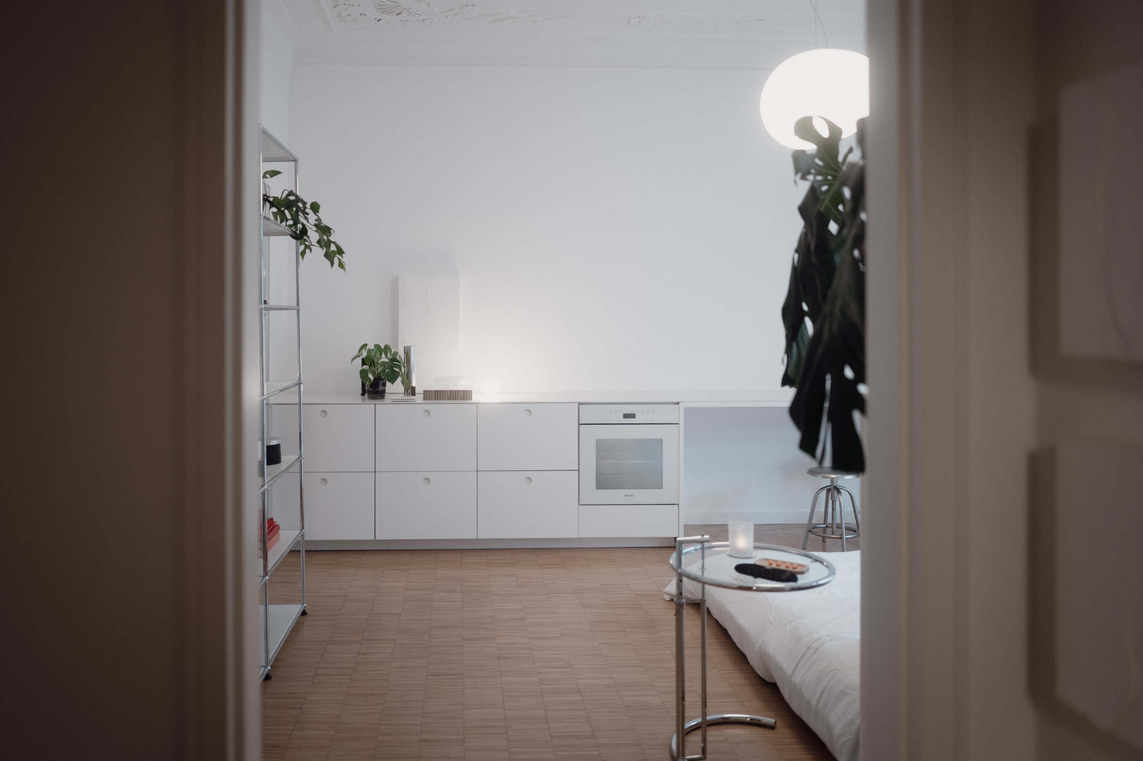 A minimalist living space features a white storage unit, a small table, and a simple bed, with a plant and pendant light enhancing the decor.