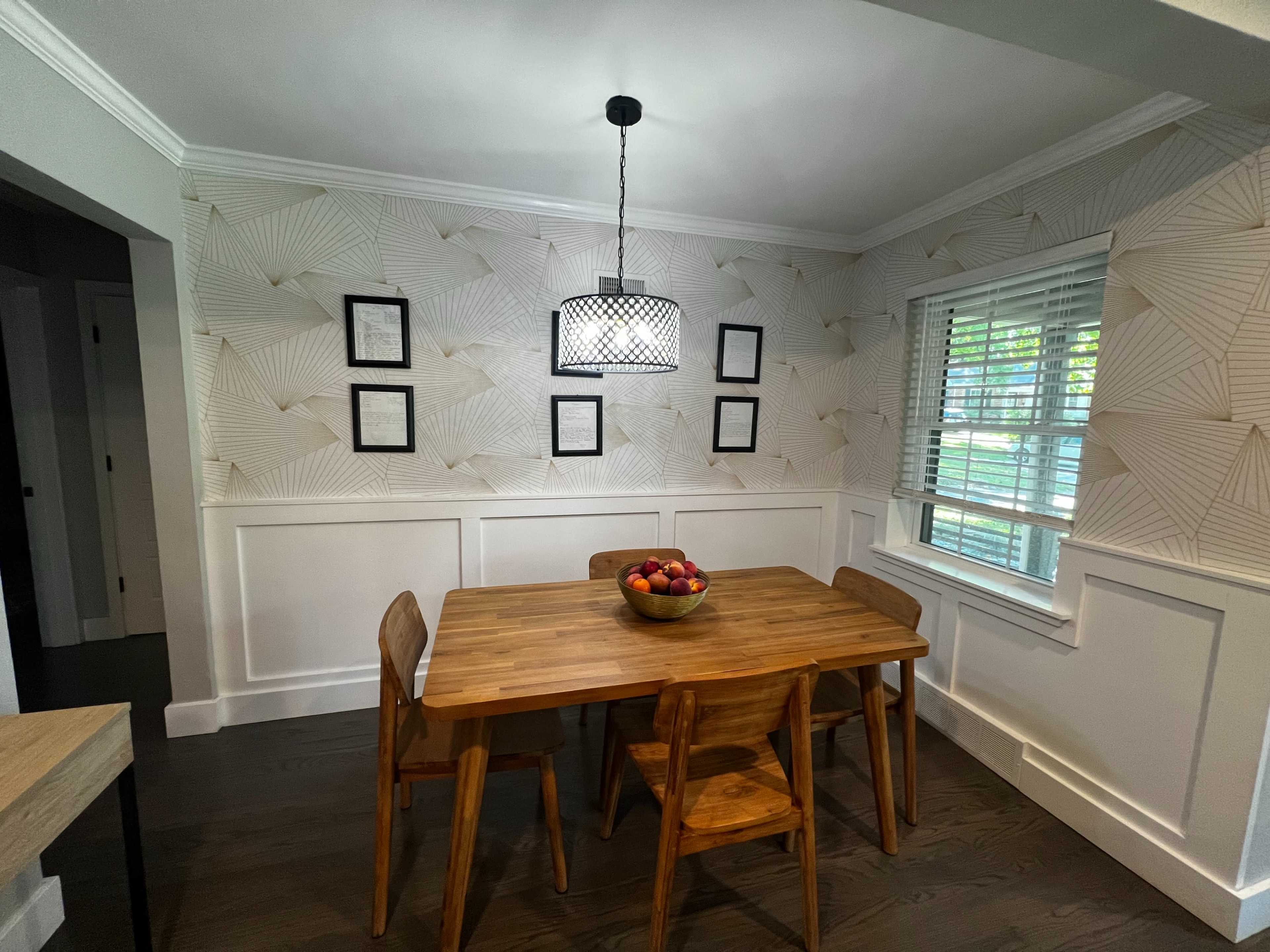 A dining area features a wooden table with a bowl of fruit, surrounded by four chairs, and has patterned wallpaper with framed pictures and a pendant light overhead.