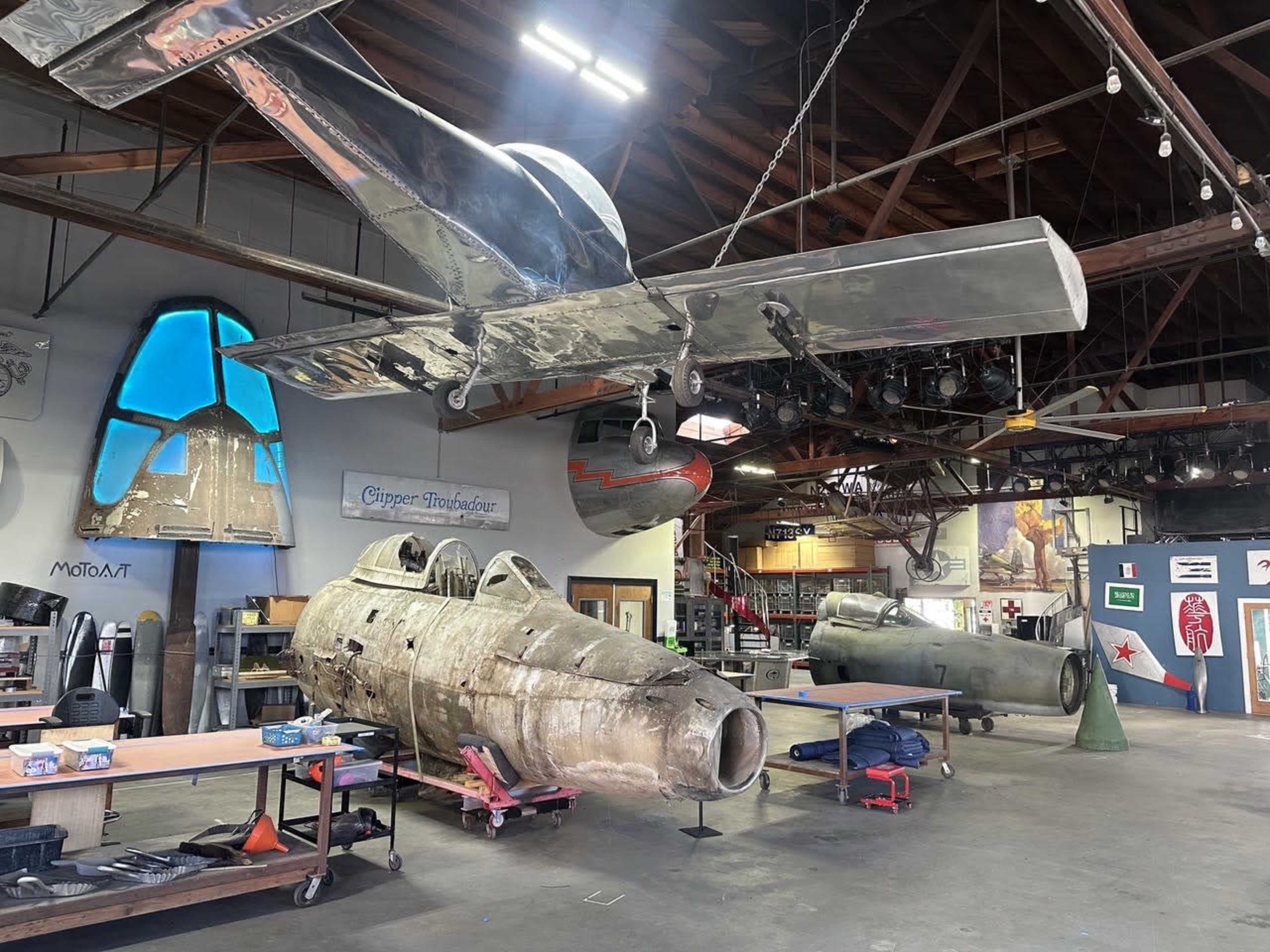 The image shows a workshop with two vintage aircraft fuselages and a large silver aircraft structure hanging from the ceiling.