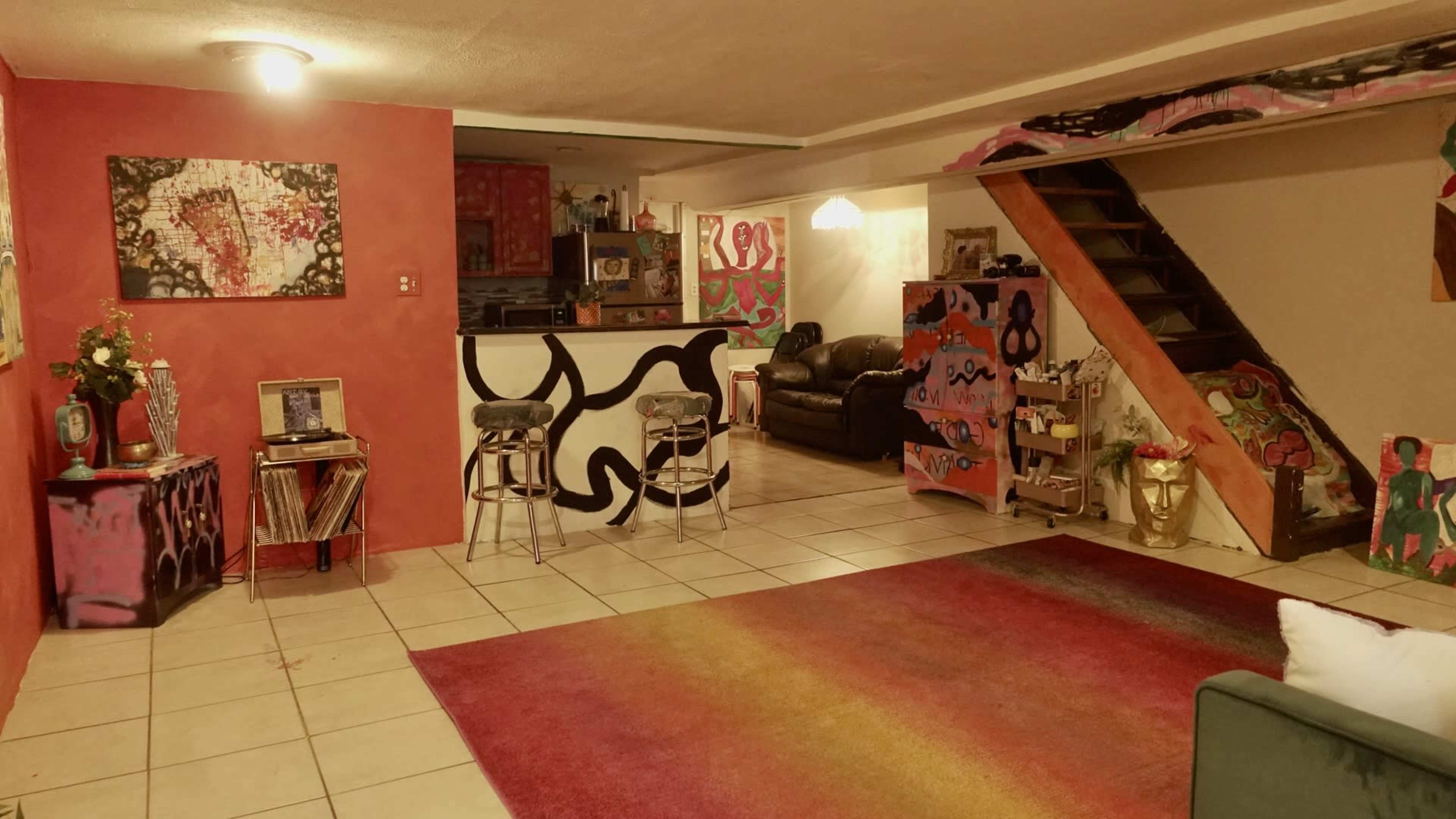 A colorful living room with abstract artwork on the walls, a staircase in the background, and a bar area with two stools.