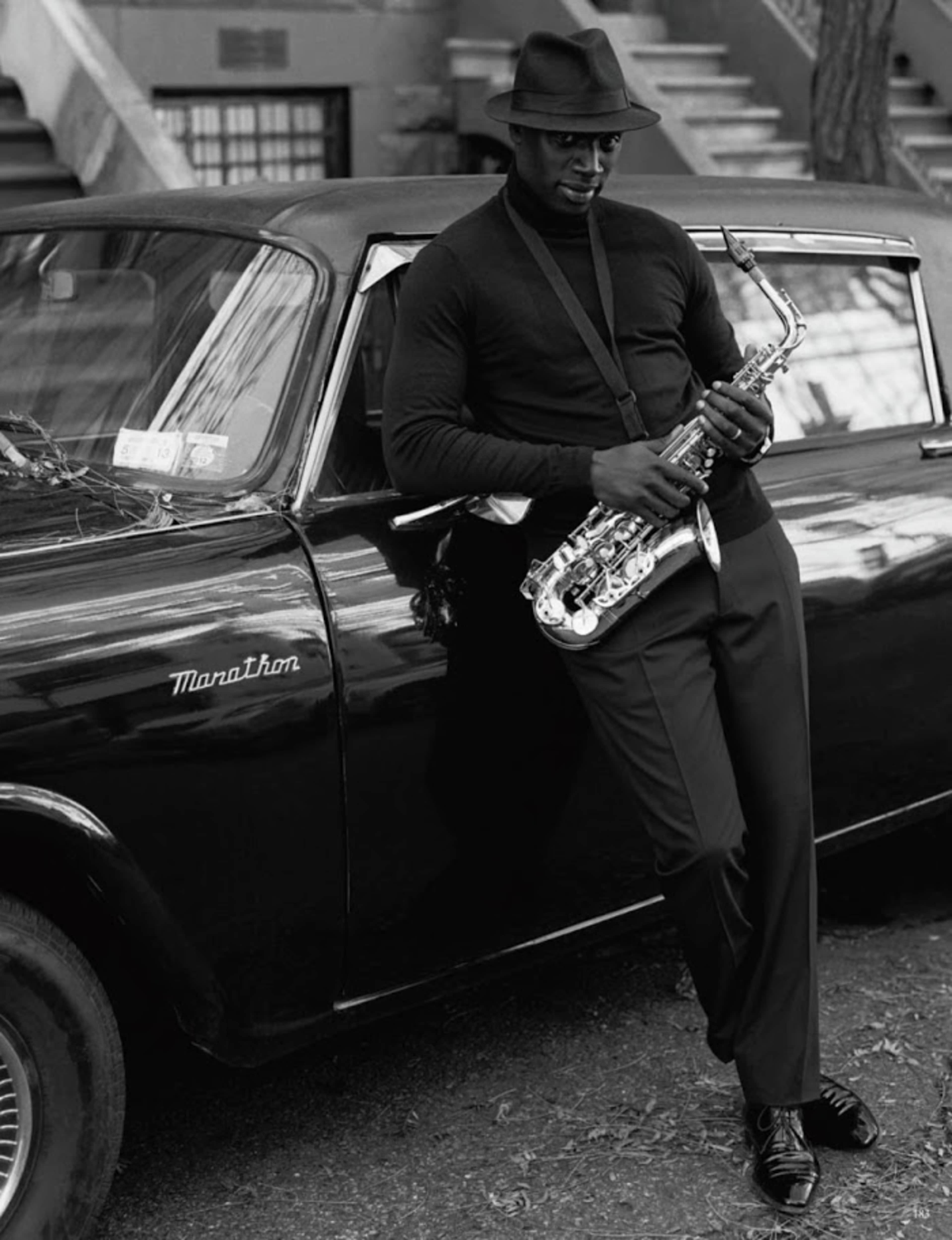 A man in a black hat leans against a vintage black car while holding a saxophone.