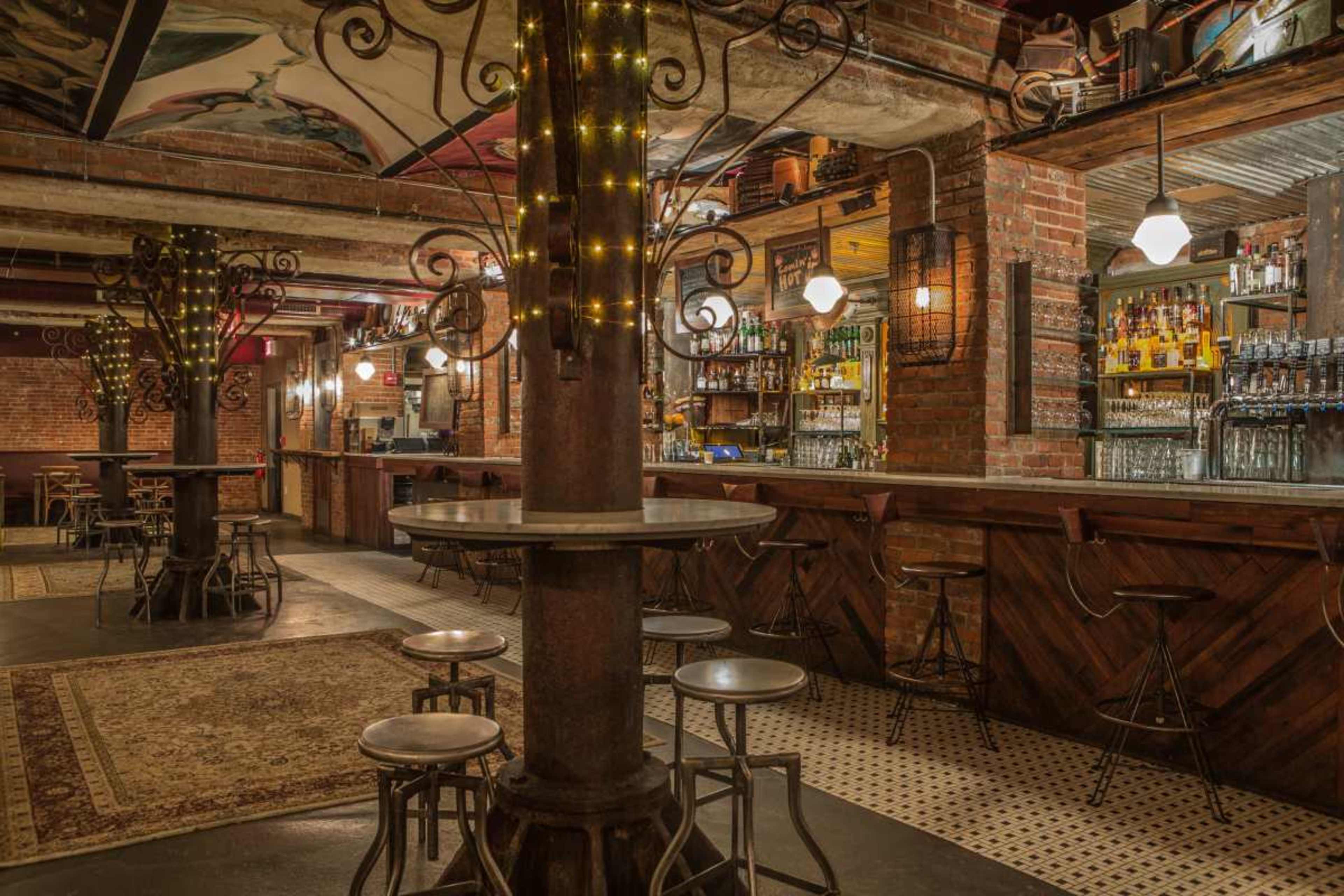 The image shows a rustic bar interior with exposed brick walls, wooden accents, a decorative column with lights, and several high-top tables surrounded by metal stools.