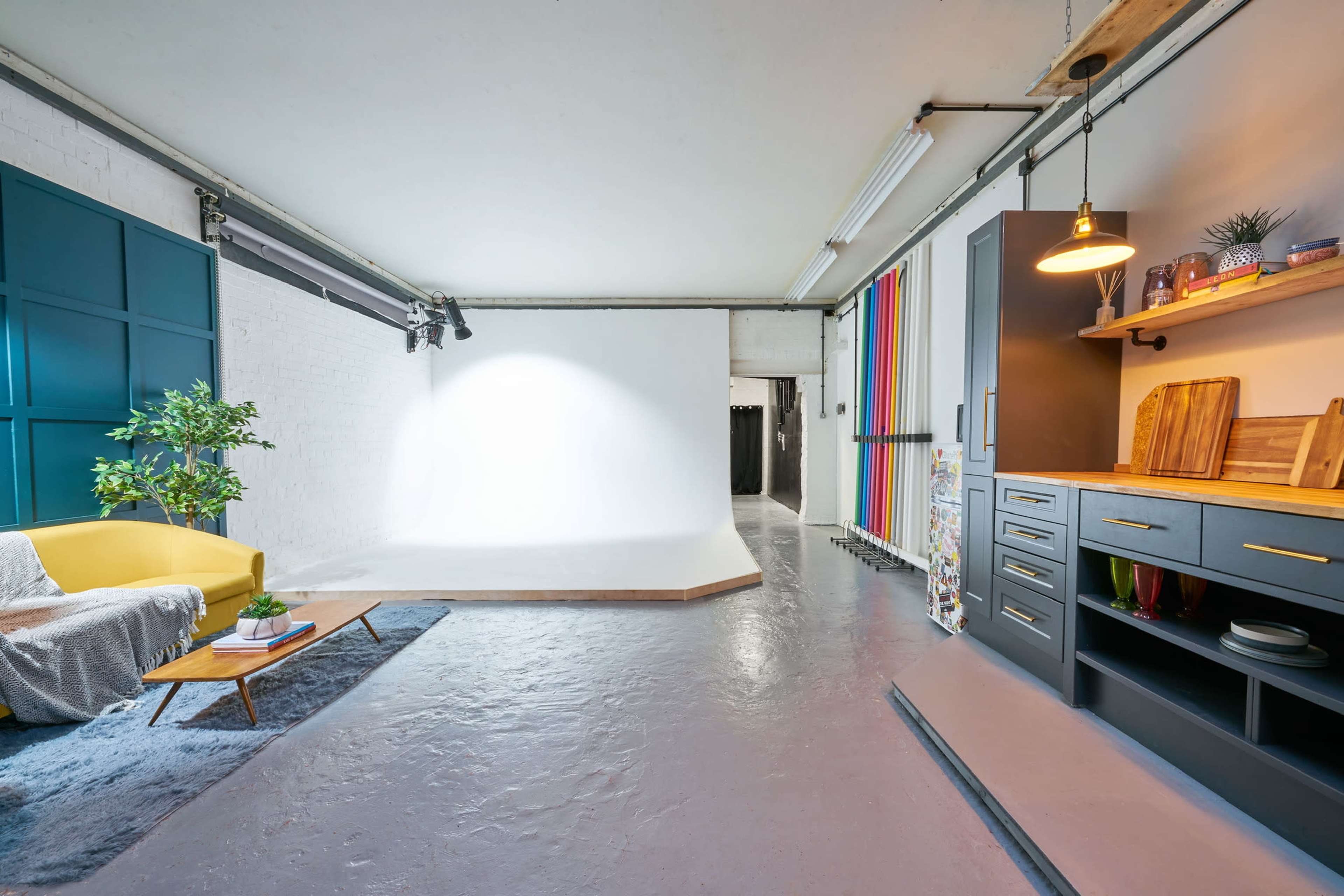 A modern studio space featuring a yellow sofa, a wooden coffee table, and a white backdrop for photography, with colorful draped panels and storage cabinets in the background.