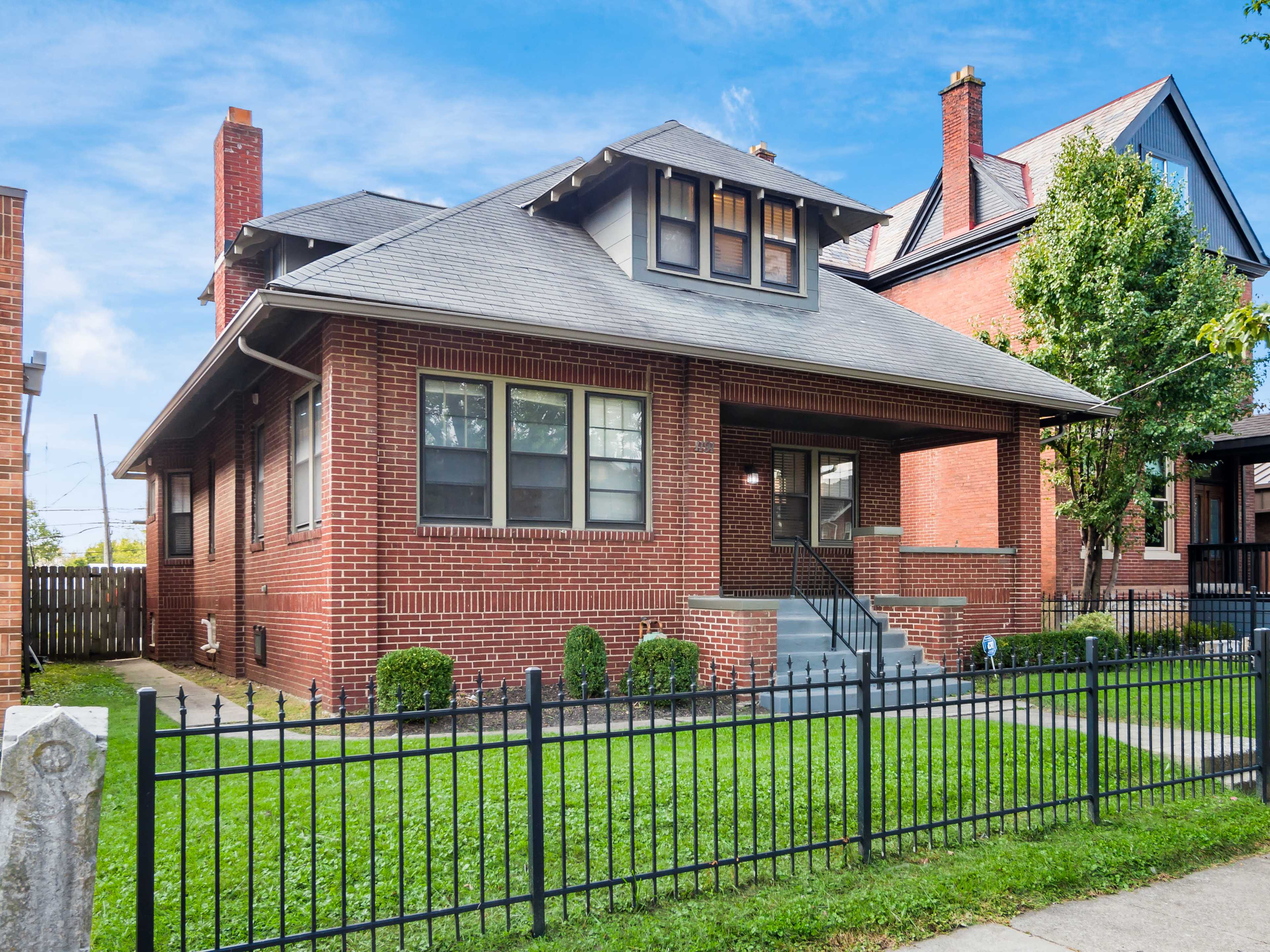A large brick bungalow with a front porch and a manicured lawn is surrounded by a black wrought-iron fence.