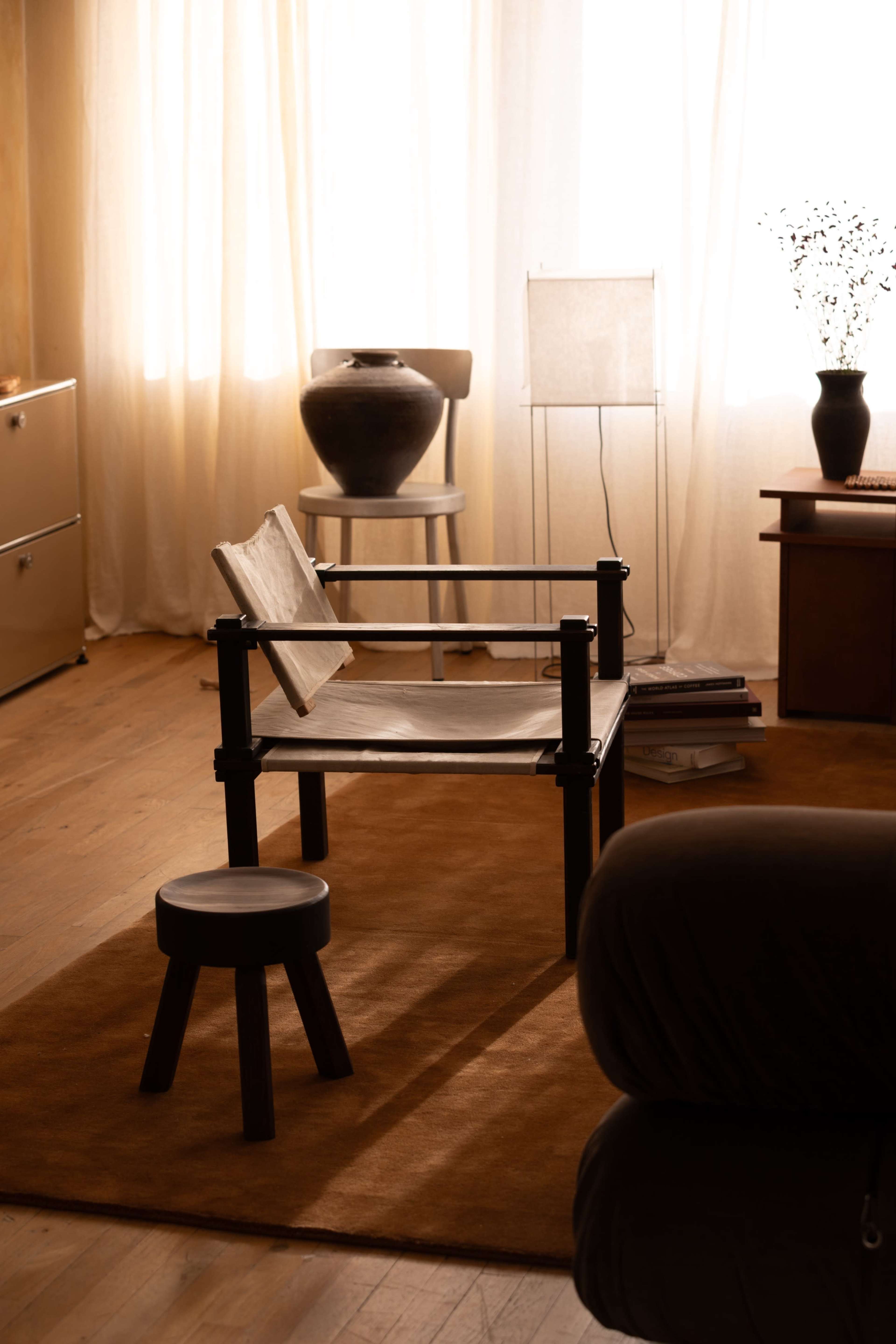 The image shows a minimalist living space featuring a light-colored chair, a small stool, a large vase, and a soft rug under warm natural light.