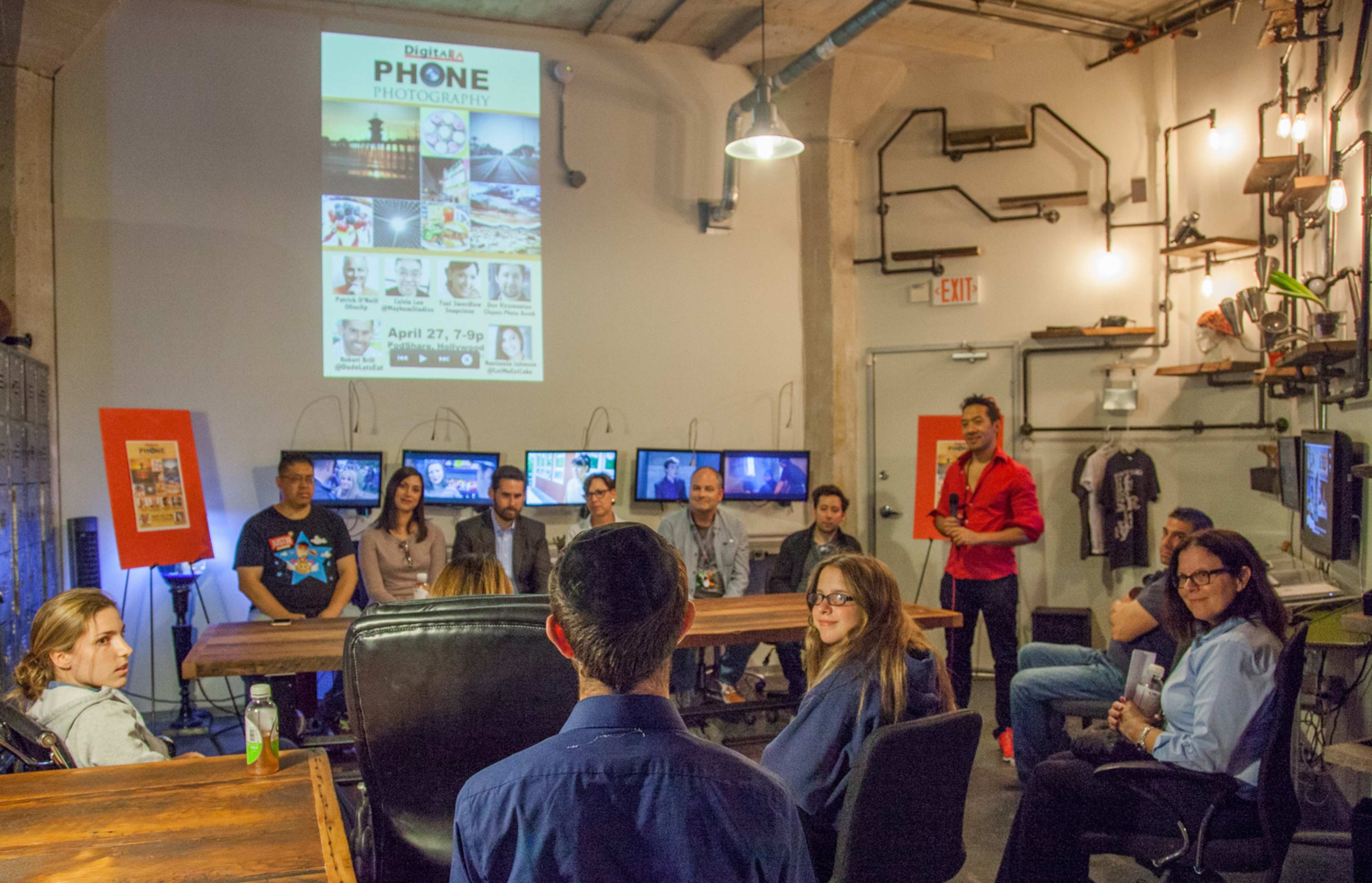 A group of people sits in a well-lit room with screens displaying photography, while a presenter stands in front of a large poster announcing a phone photography event.