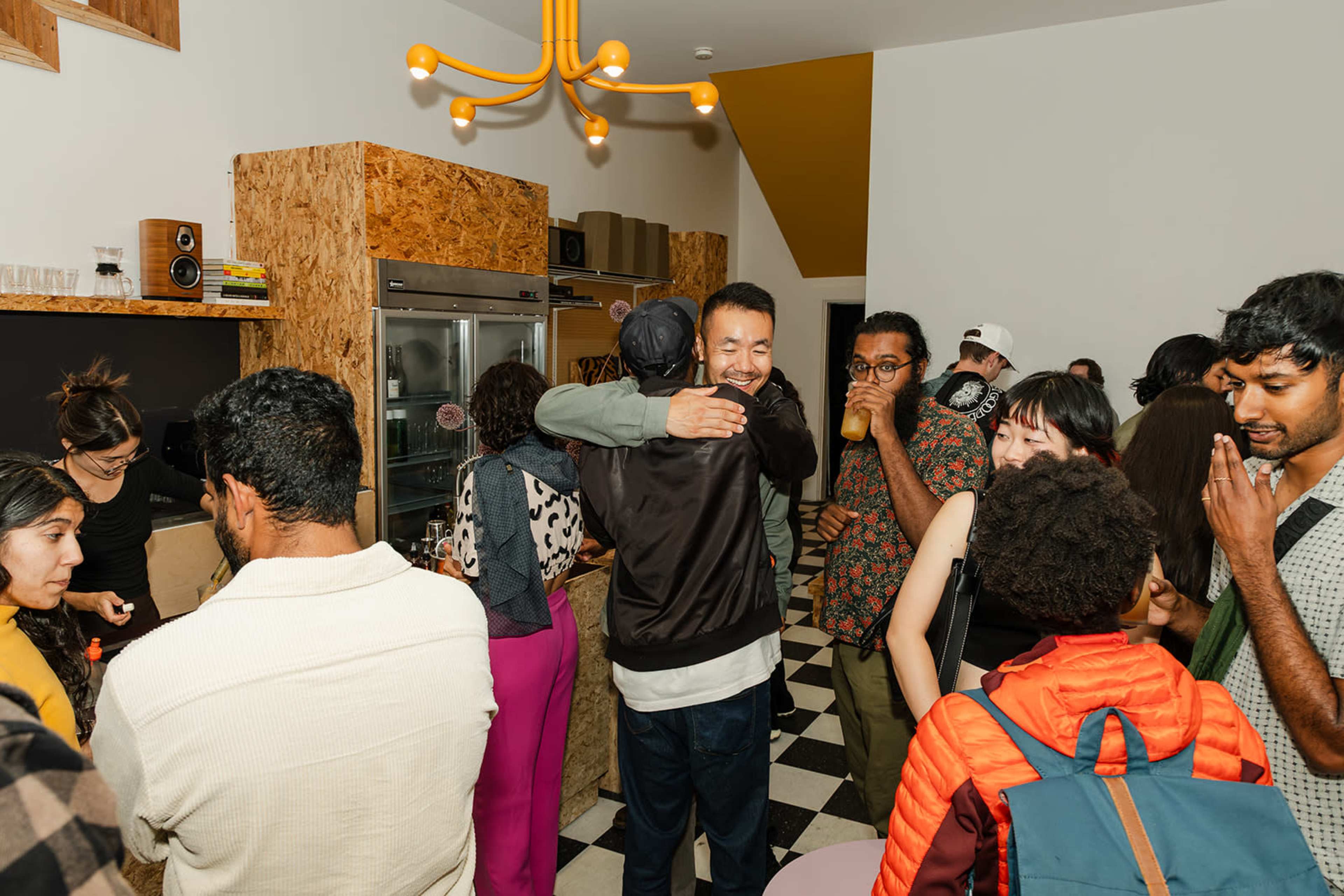 A group of people mingle and socialize in a cozy, modern indoor space with hardwood accents and a checkered floor.