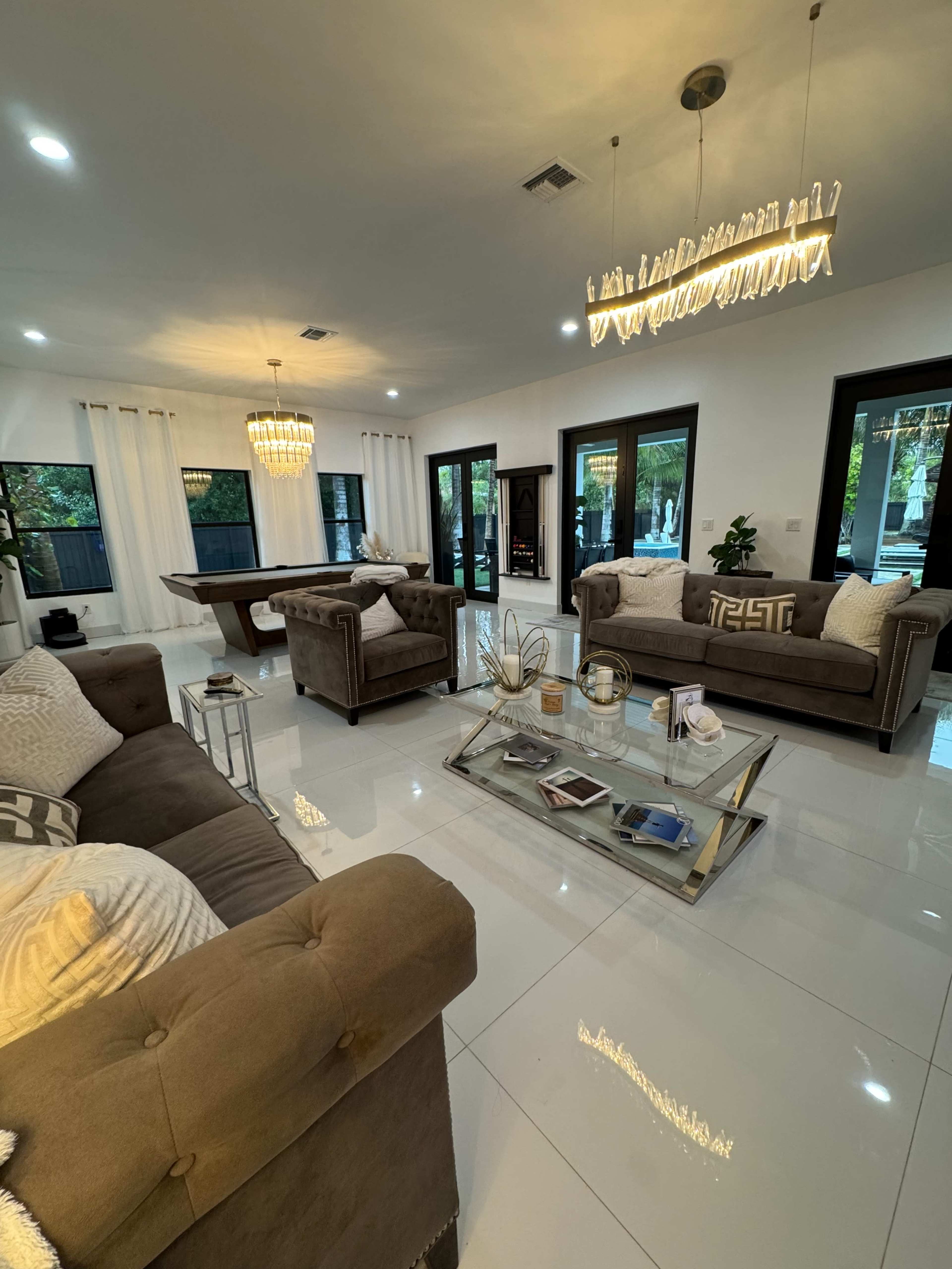 The image shows a modern living room featuring two brown sofas, a glass coffee table, and a pool table, with large windows allowing natural light to fill the space.