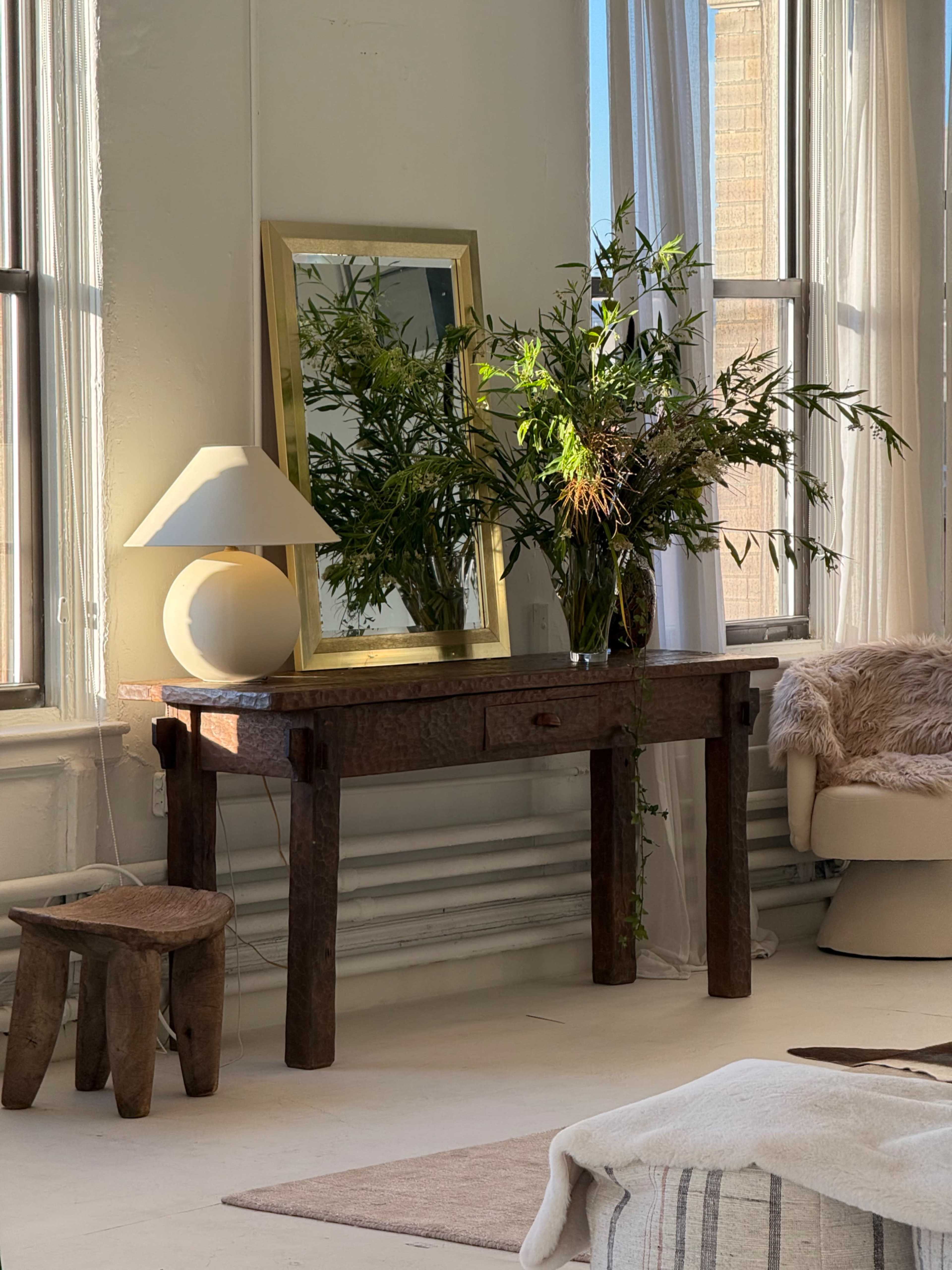 The scene features a wooden console table with a lamp and a large mirror, surrounded by natural light and decorative plants.