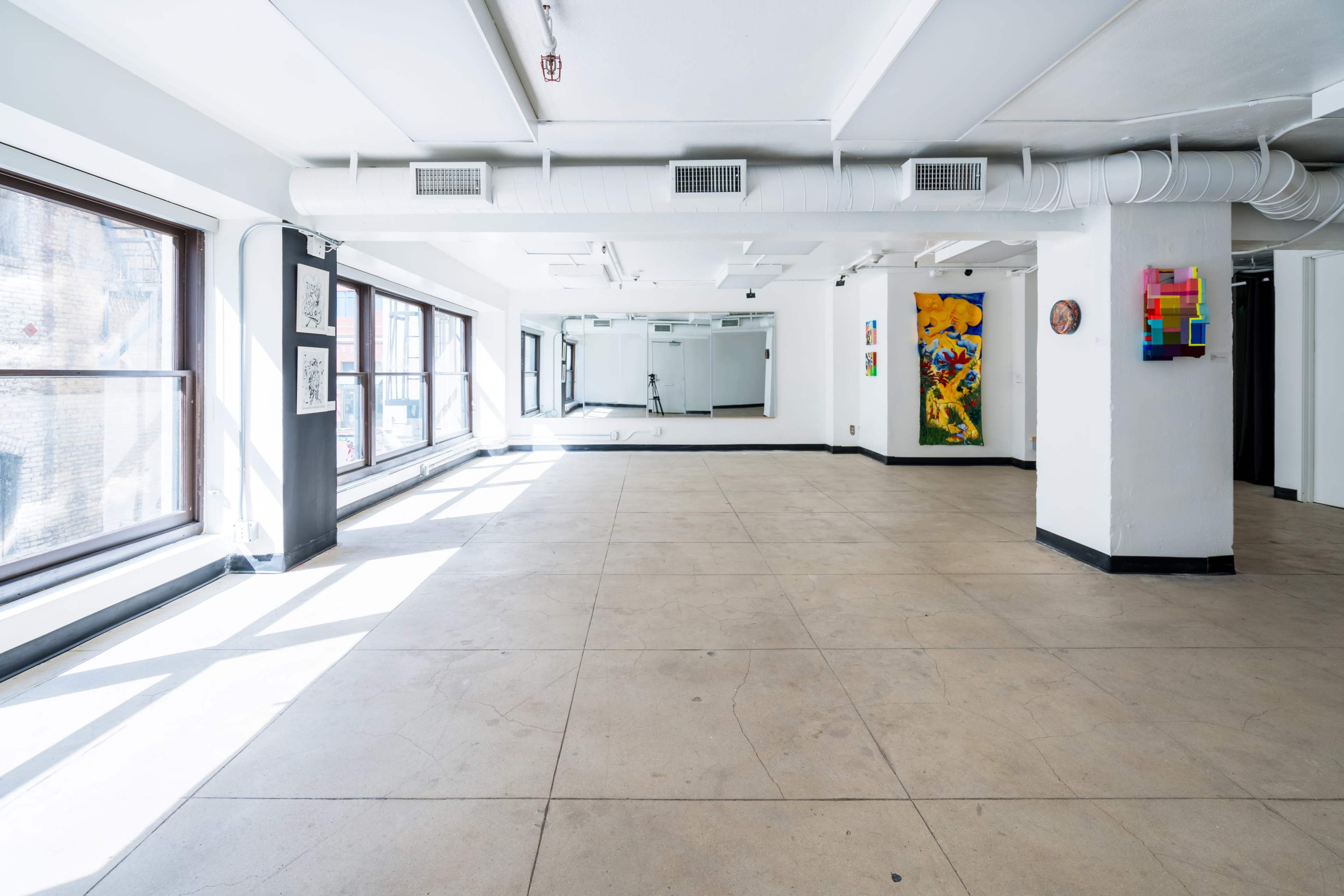 A spacious, empty gallery with large windows and white walls, featuring colorful artwork on two sides.