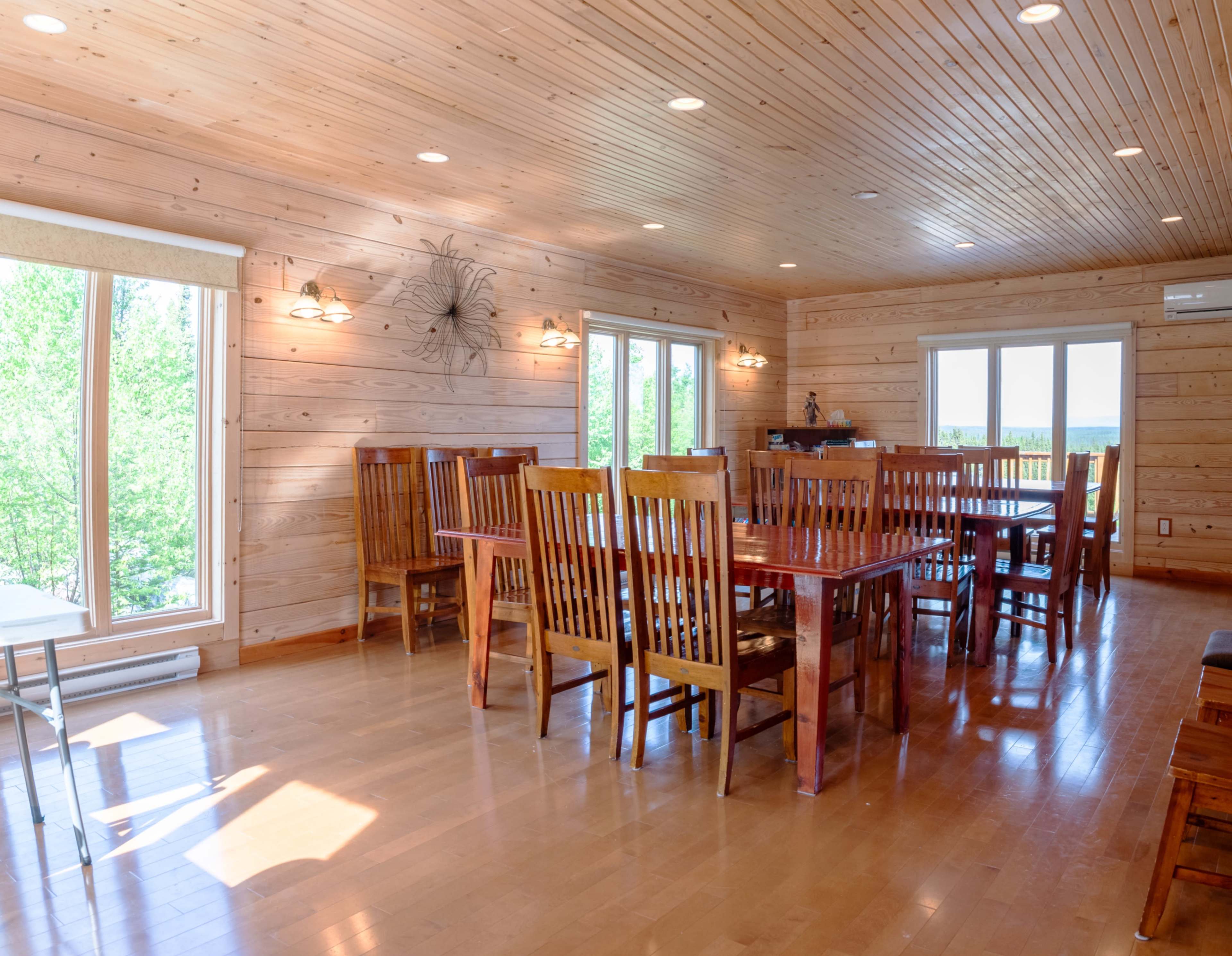 A spacious dining area features wooden walls and a large table surrounded by multiple wooden chairs, illuminated by natural light from large windows.