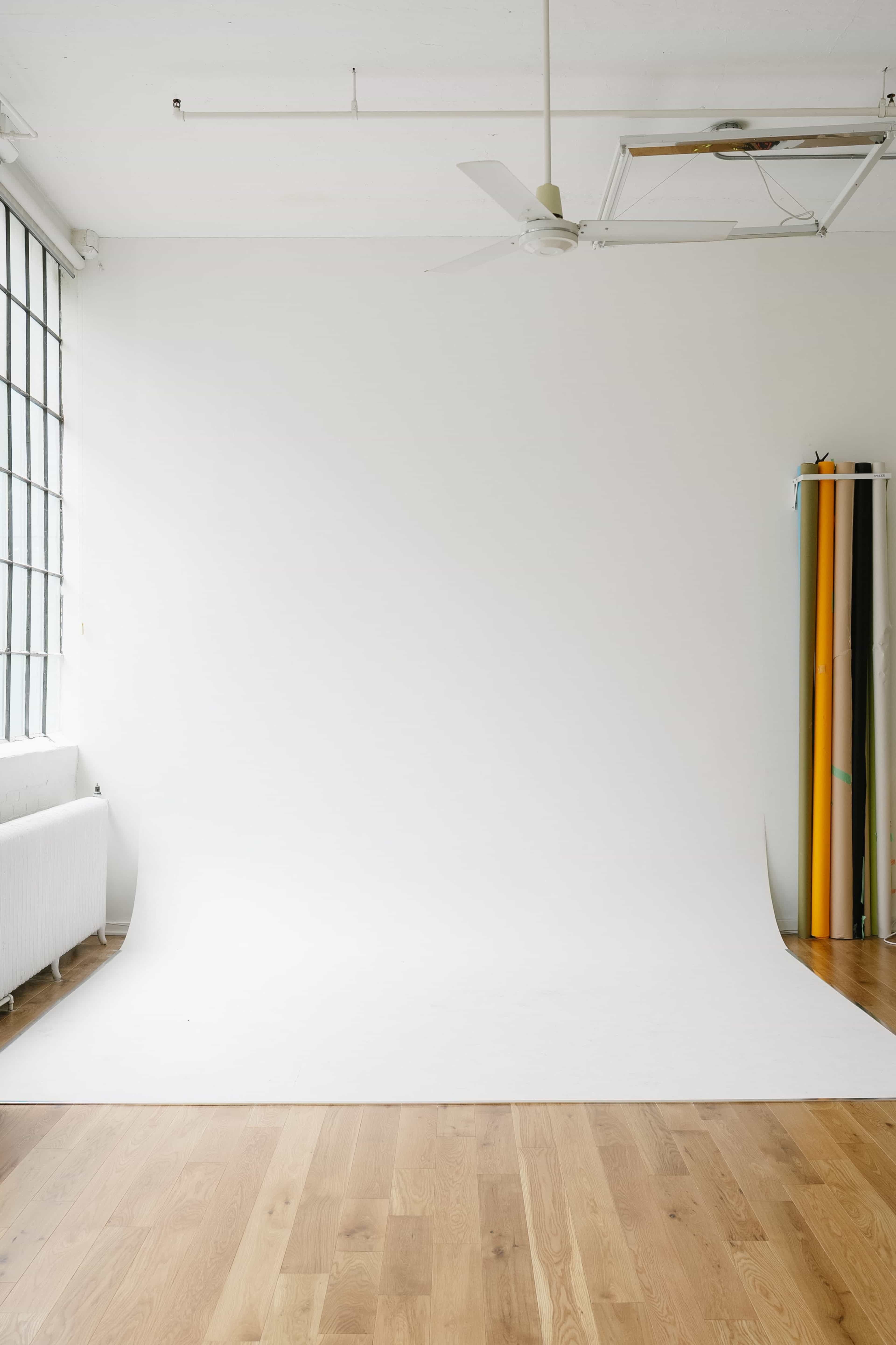 The image shows a spacious, bright studio with a large white backdrop, wooden flooring, and a corner containing rolls of colored backdrops.