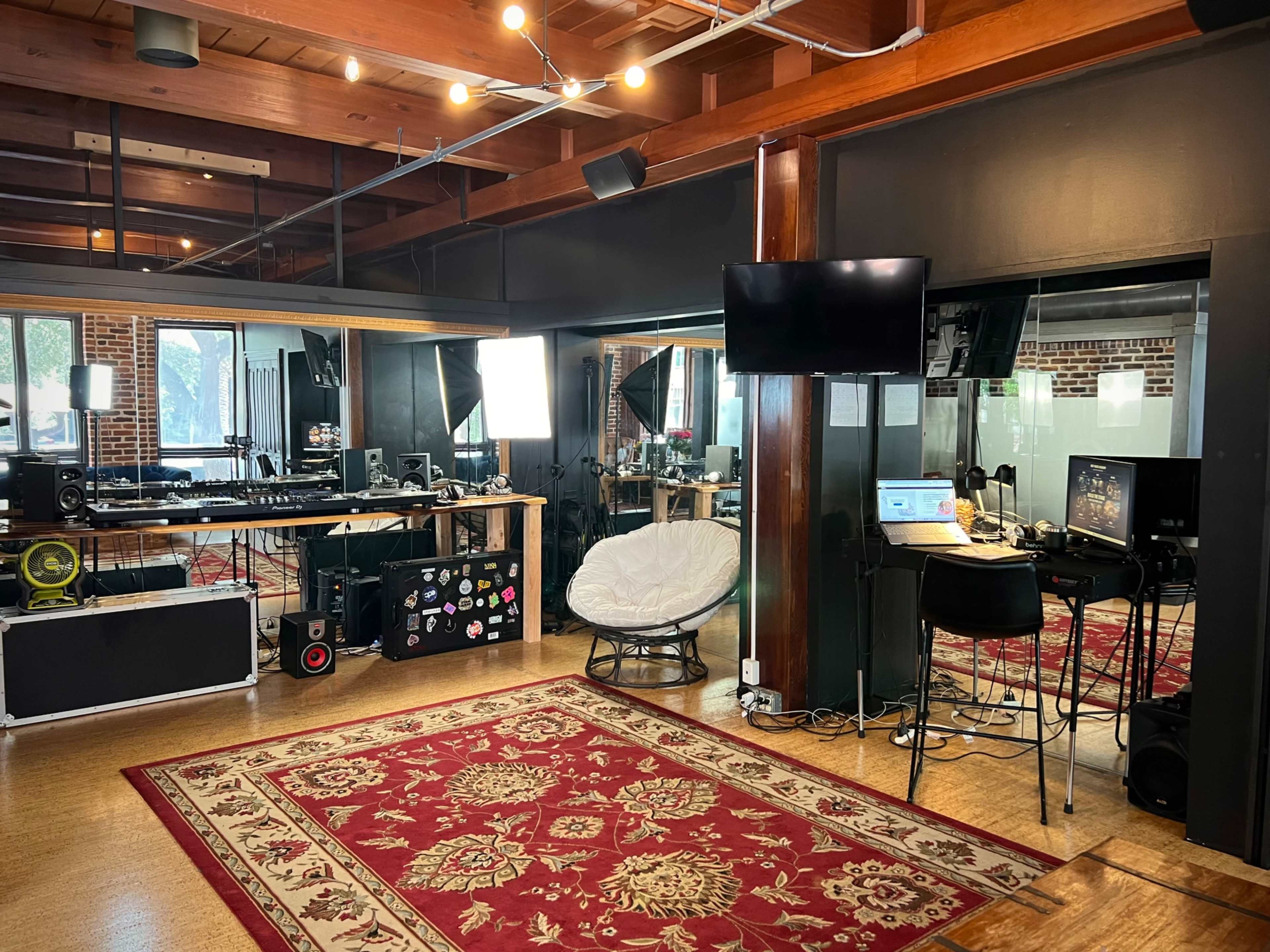 The image shows a spacious studio with wooden beams, a red patterned rug, a seating area, and multiple music production equipment setups.