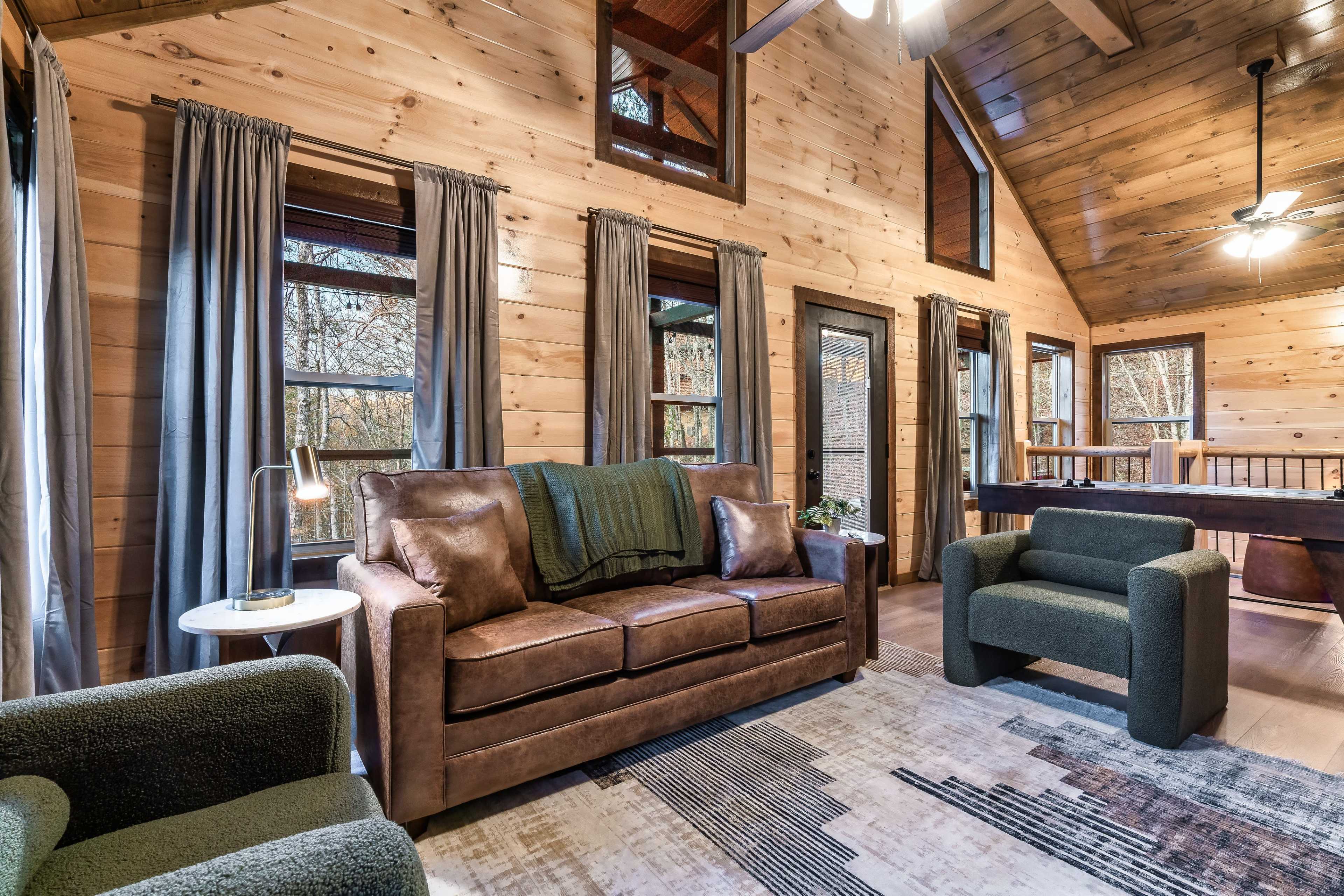 The image shows a cozy, wooden cabin living room with a brown couch, two green armchairs, and large windows letting in natural light.