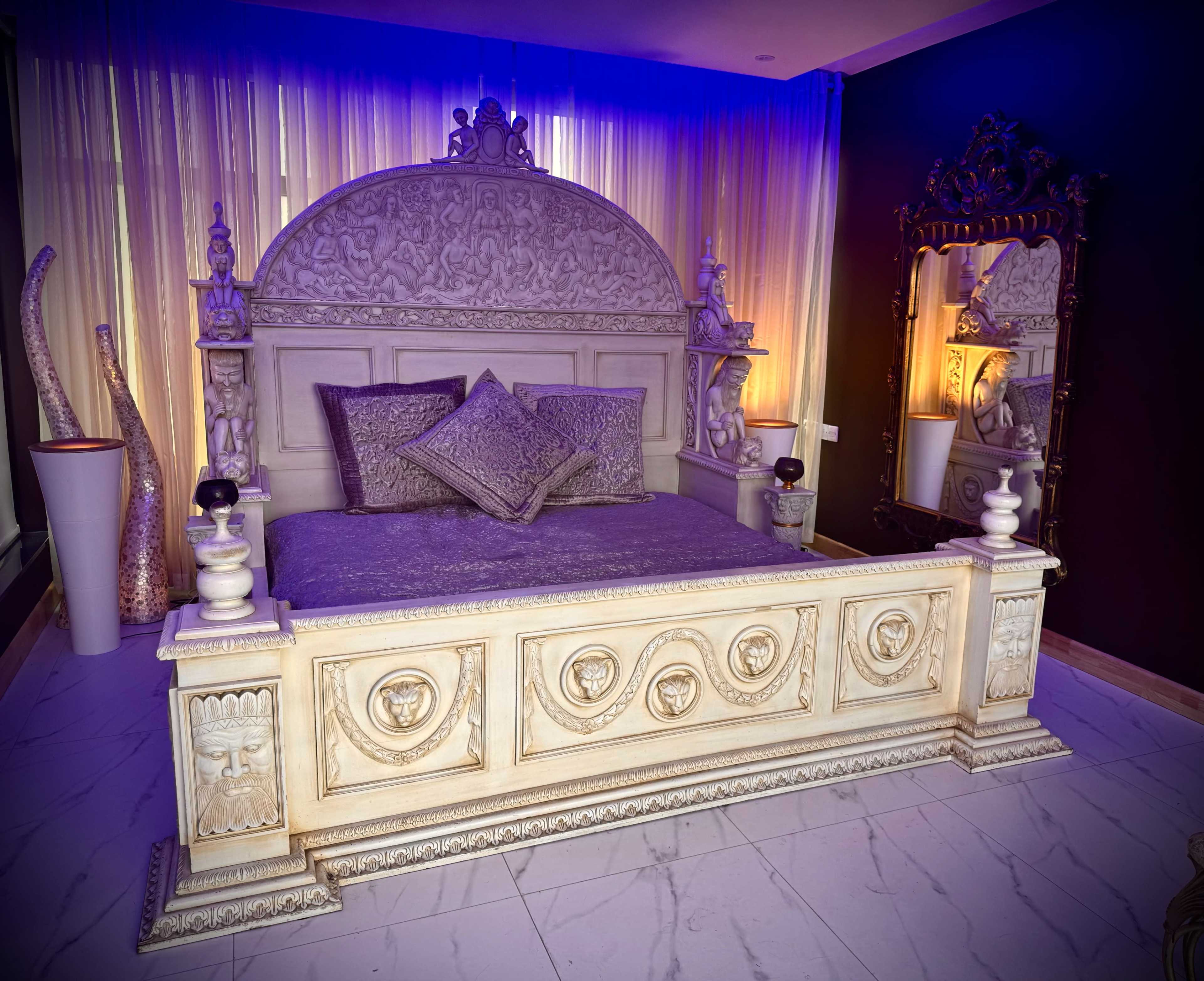 An ornate, carved bed with decorative pillows in a dimly lit room featuring elegant drapery and a mirror.
