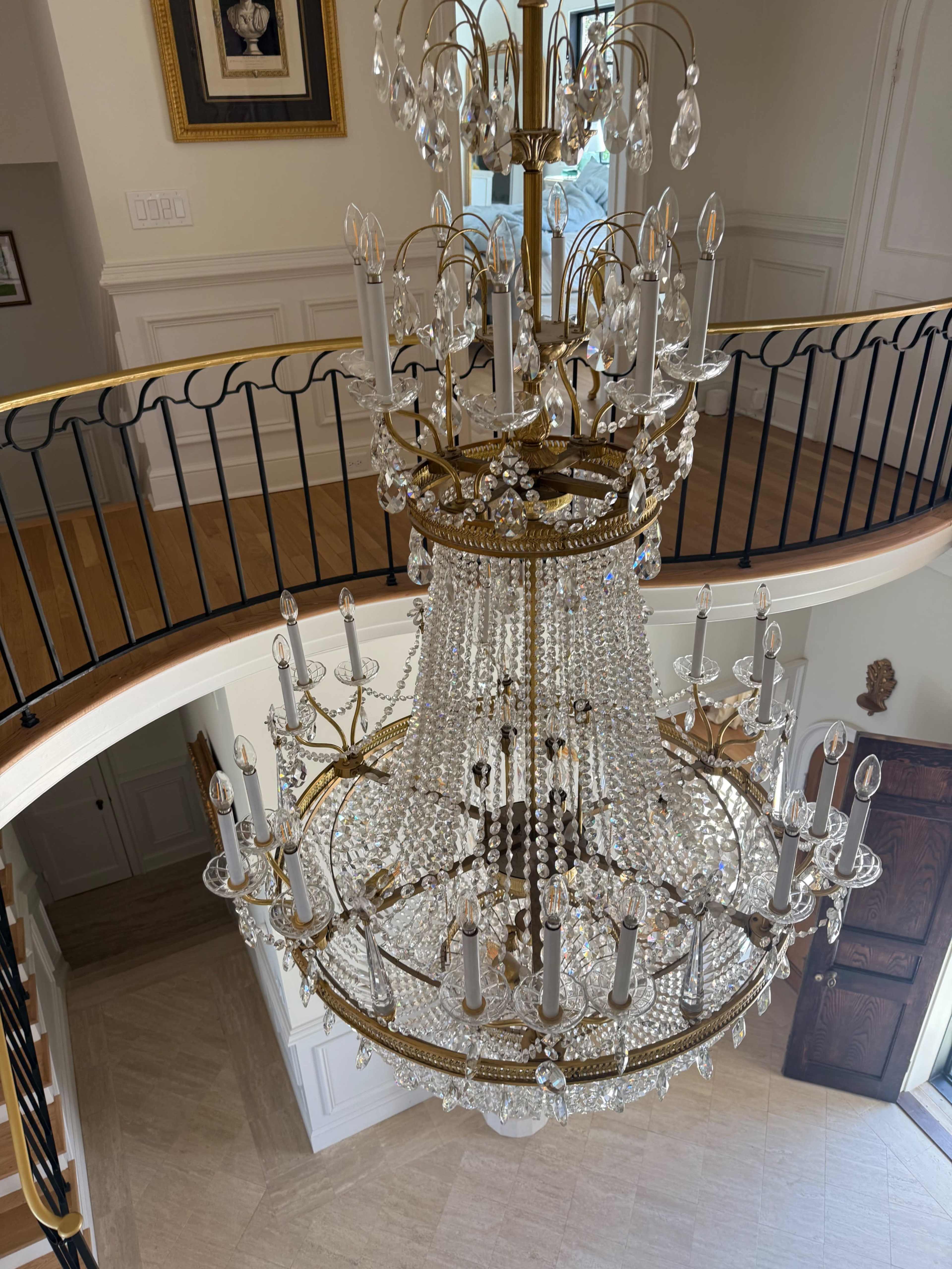 A large crystal chandelier hangs from the ceiling of an ornate staircase, surrounded by elegant railings and decorative walls.
