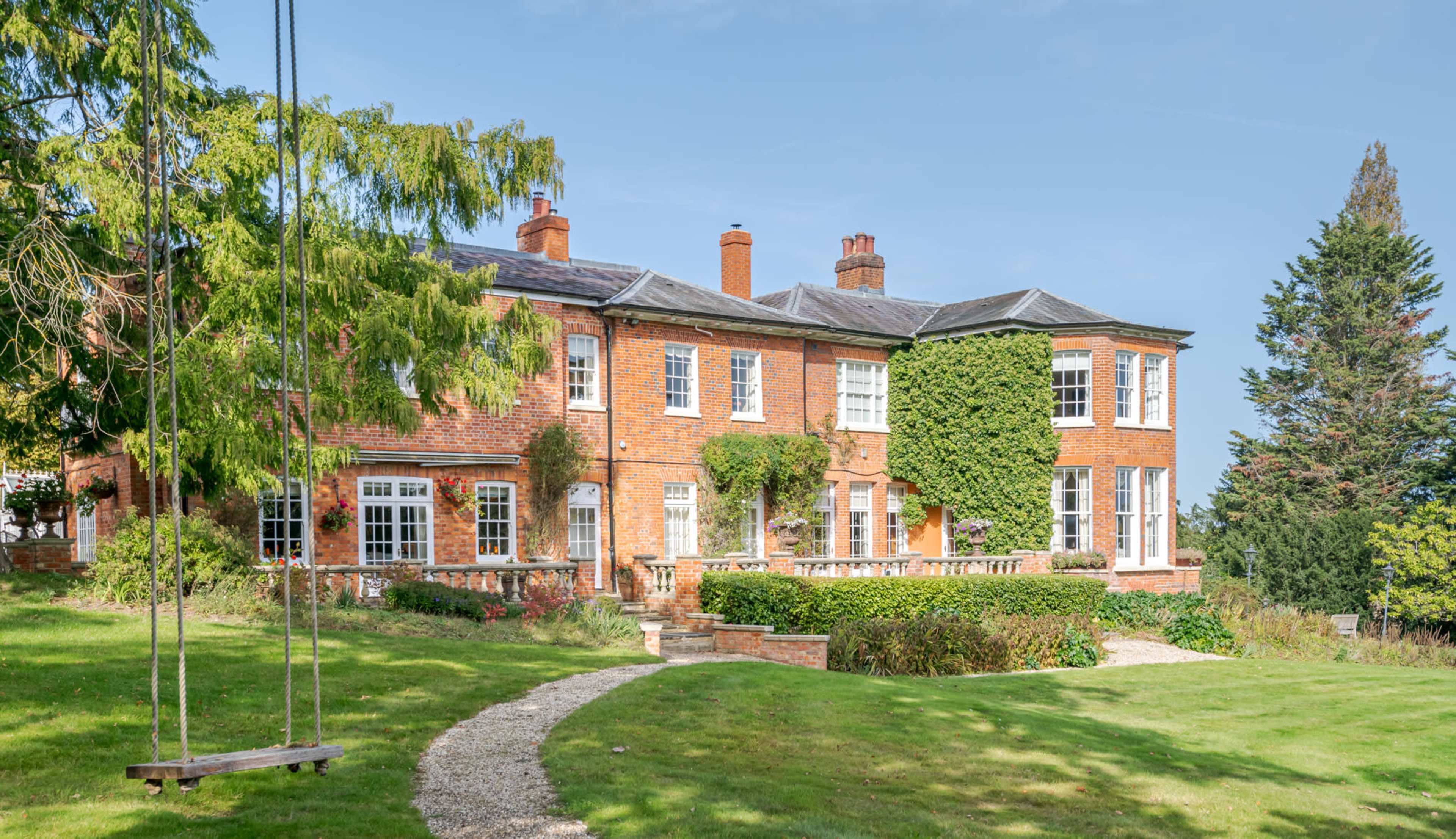 A large brick house with multiple windows is situated on a lush green lawn, framed by trees and a stone pathway.
