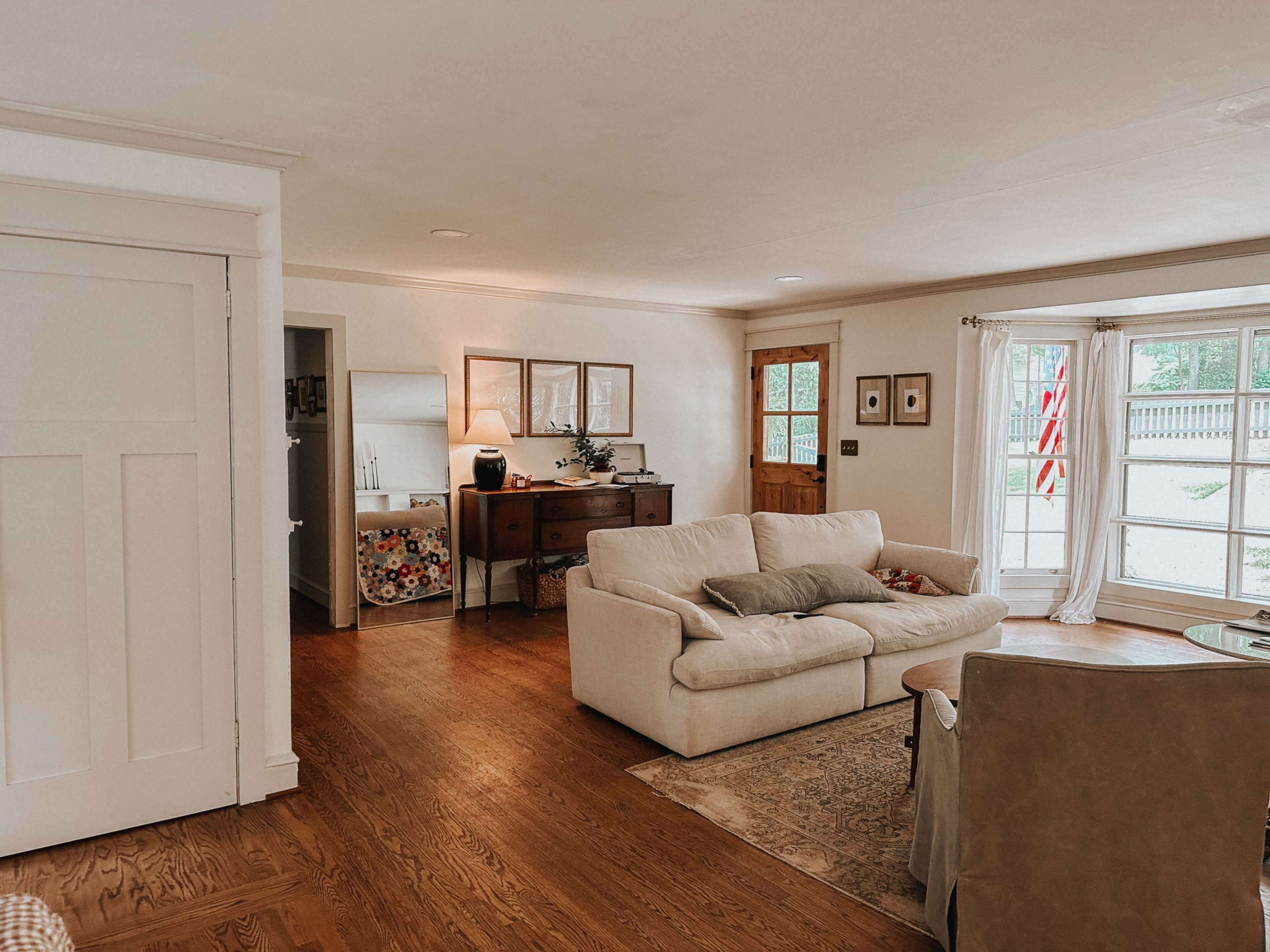 The living room features a cream-colored sofa, a wooden sideboard, and large windows displaying a view of the outdoors.