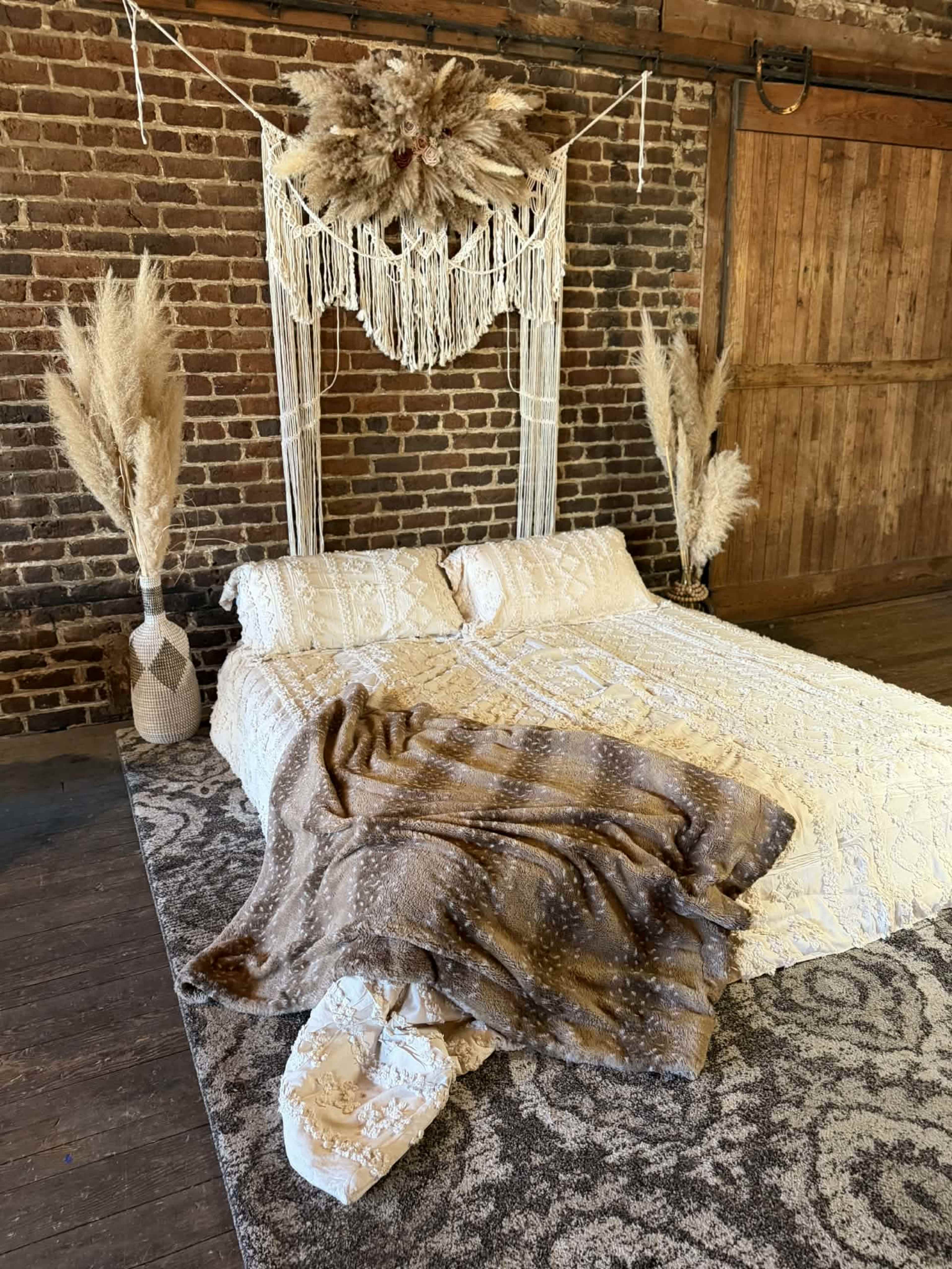 The image shows a bed covered with textured bedding and a patterned throw, flanked by decorative plants and a macramé wall hanging against a brick wall.