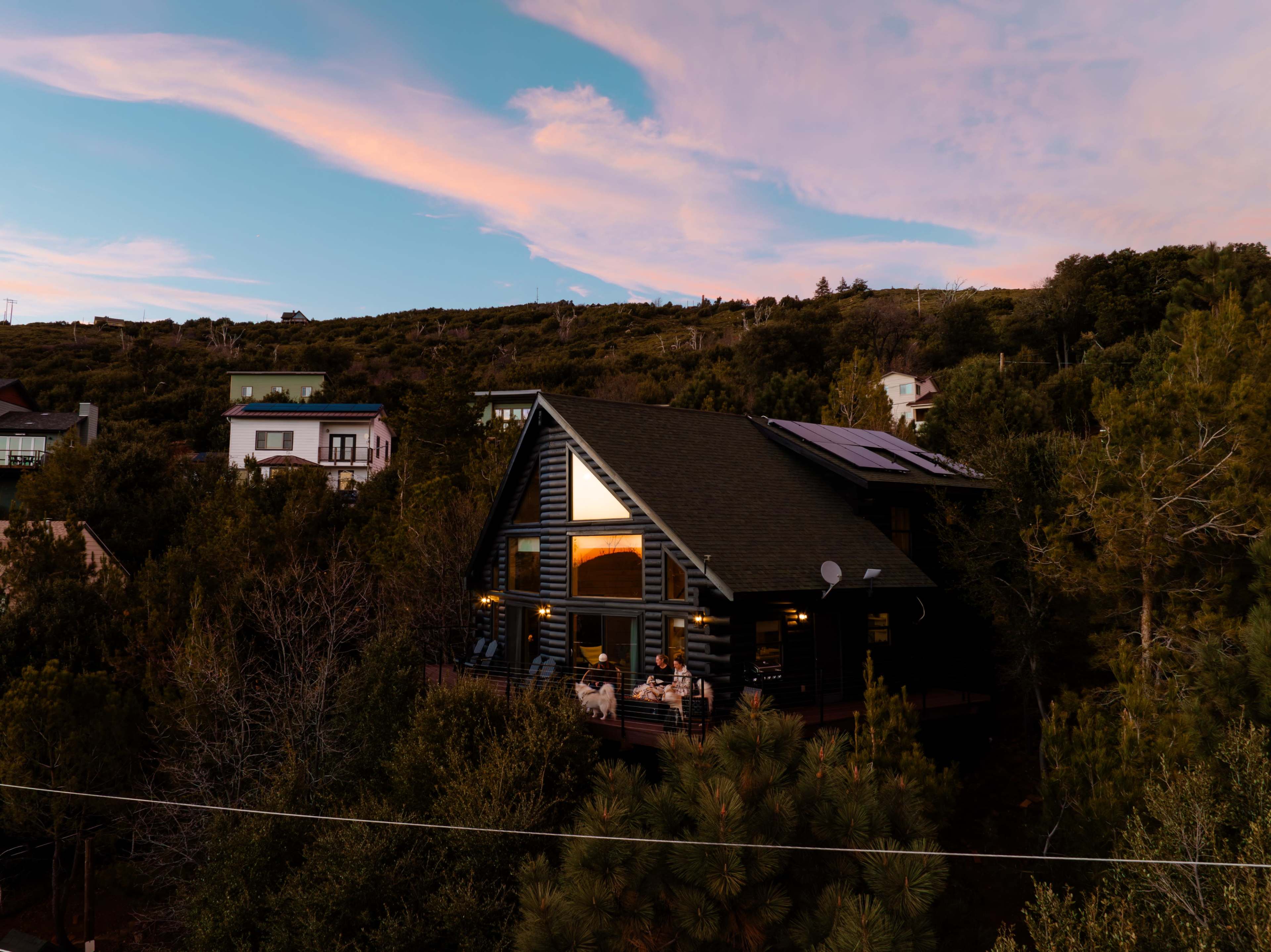 A dark wooden house with large windows sits among greenery, with people visible on the deck during sunset.