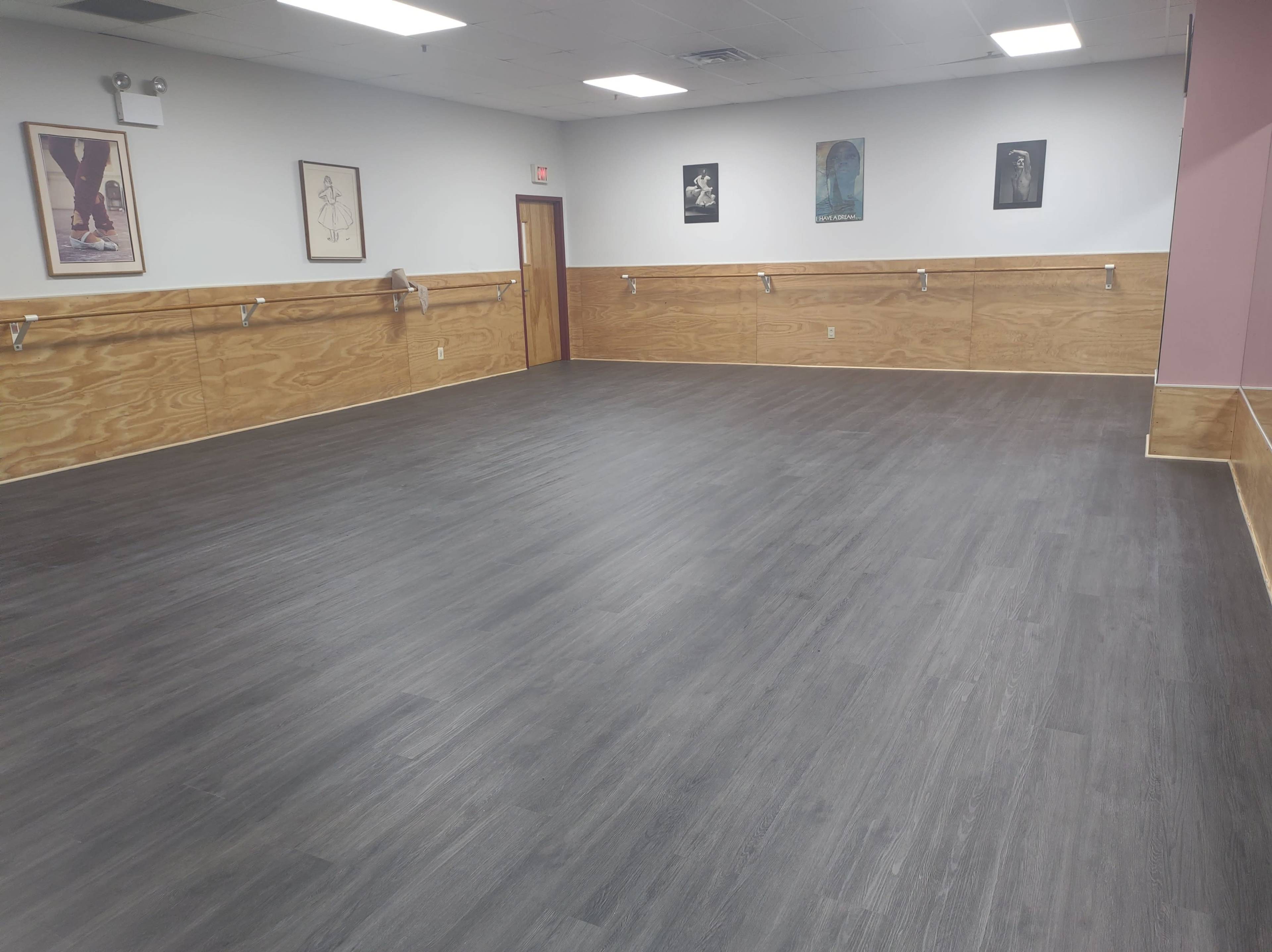 The image shows an empty room with hardwood flooring and wooden wall paneling, featuring framed artworks on the walls.