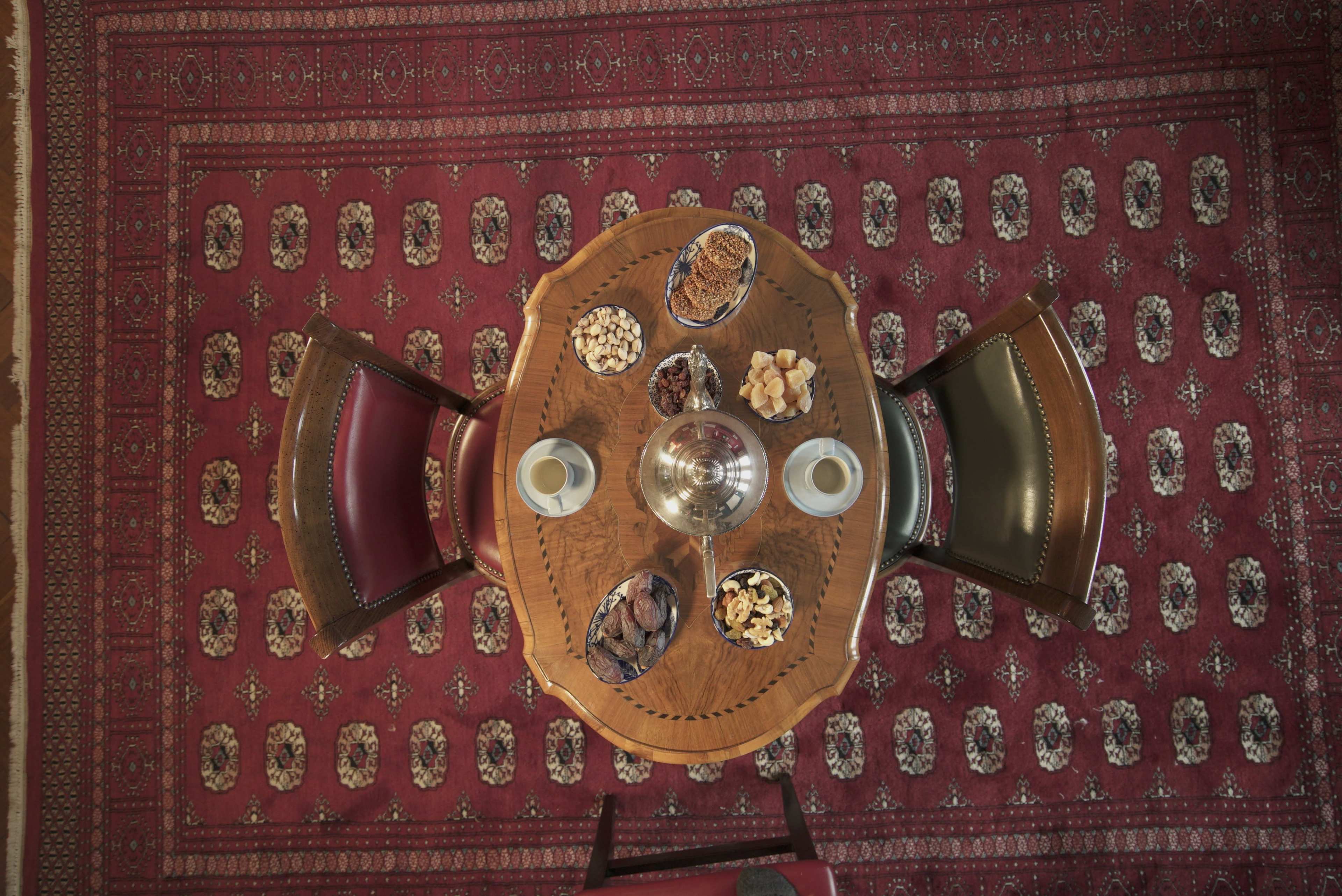 A round wooden table is set with various snacks and two cups, surrounded by two chairs, on a patterned red carpet.