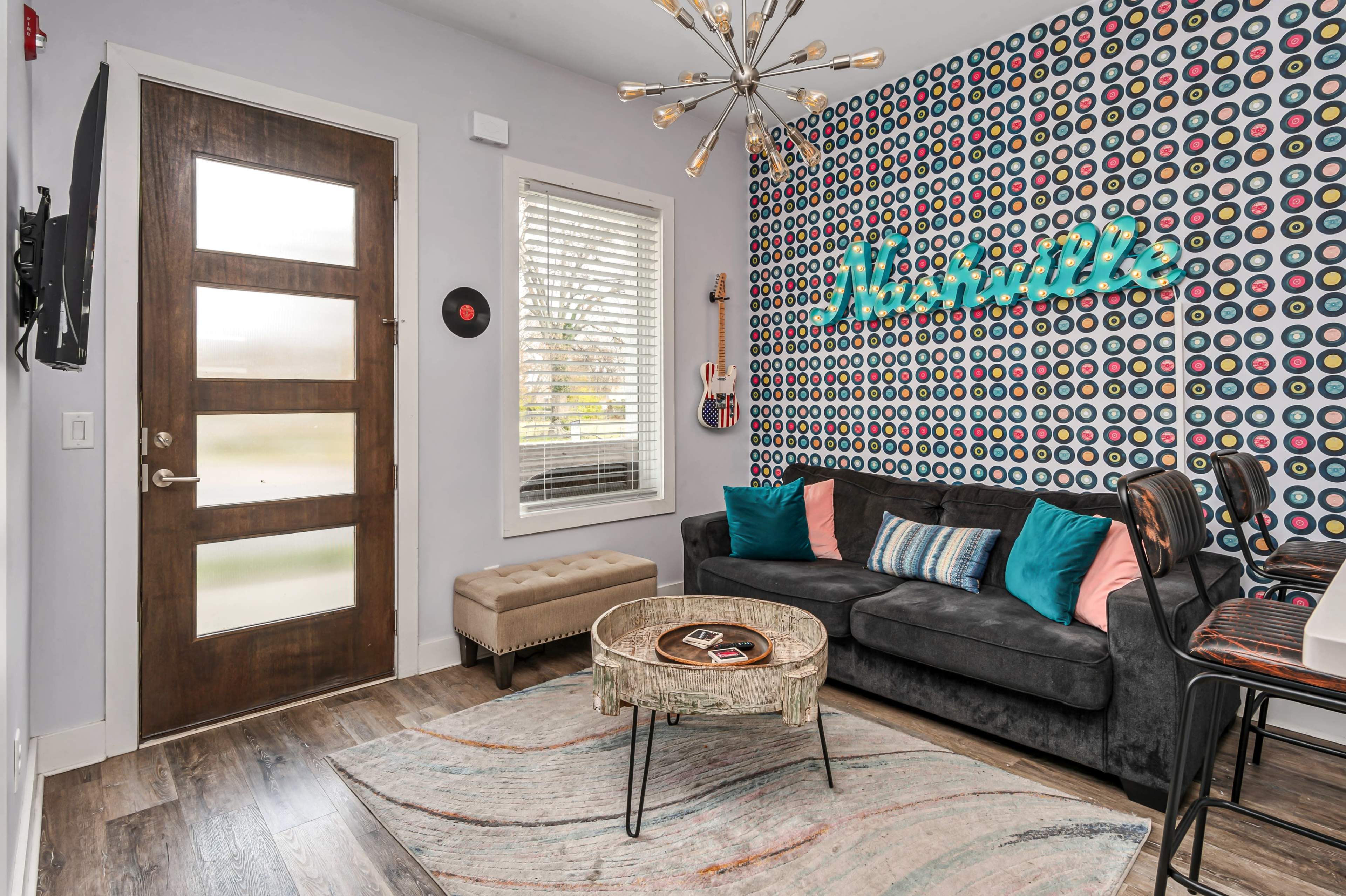 The room features a modern design with a gray sofa, colorful accent pillows, a round coffee table, and a patterned accent wall.