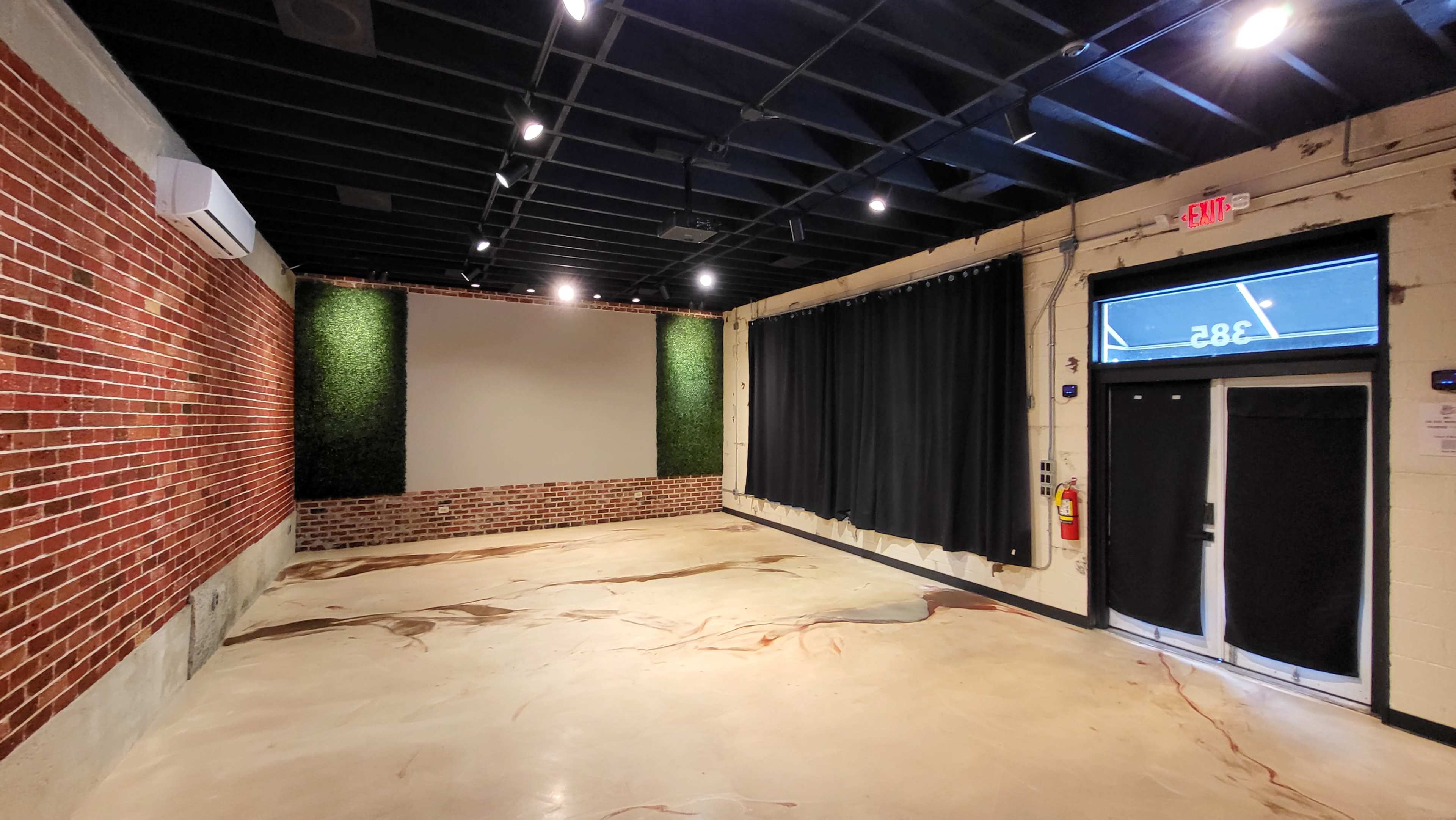 The image shows an empty room with exposed brick walls, a smooth concrete floor, a large blank screen on one side, and curtains covering a door on the other side.