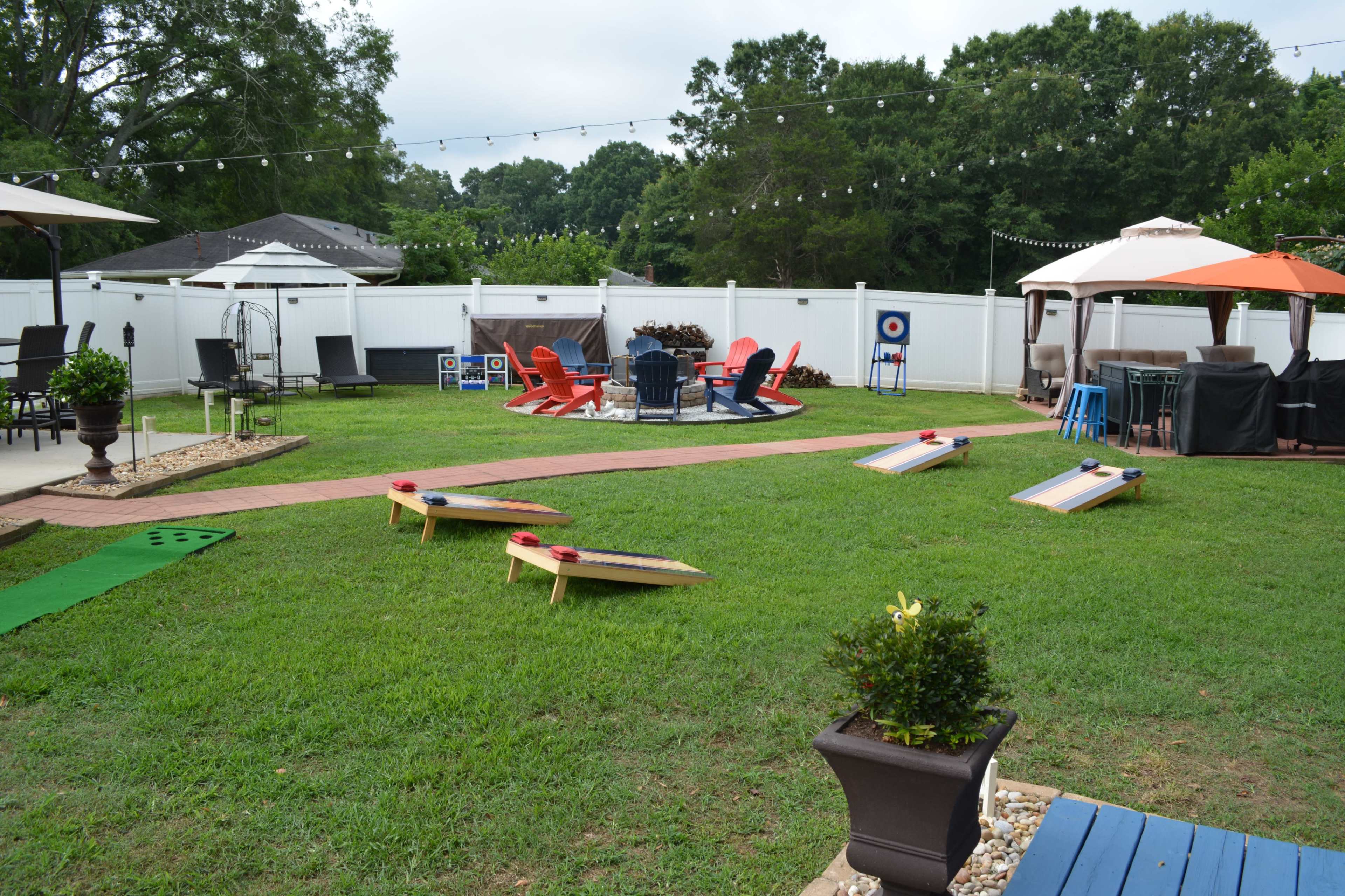 The image shows a well-maintained backyard featuring lawn games, seating areas with colorful chairs, and shade structures under a partly cloudy sky.