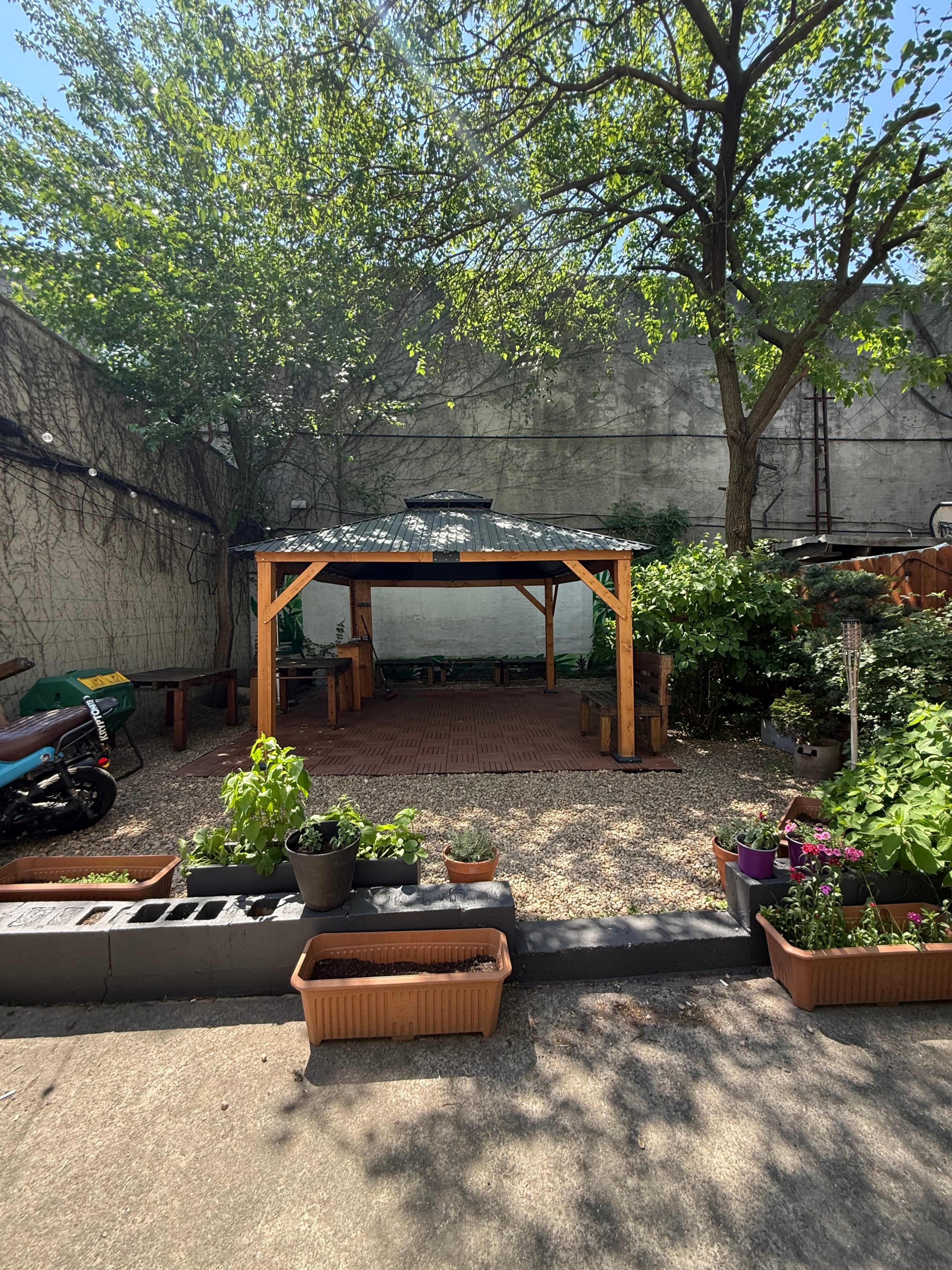 The image shows a shaded outdoor seating area with a wooden gazebo surrounded by various potted plants and a motorcycle parked to the side.