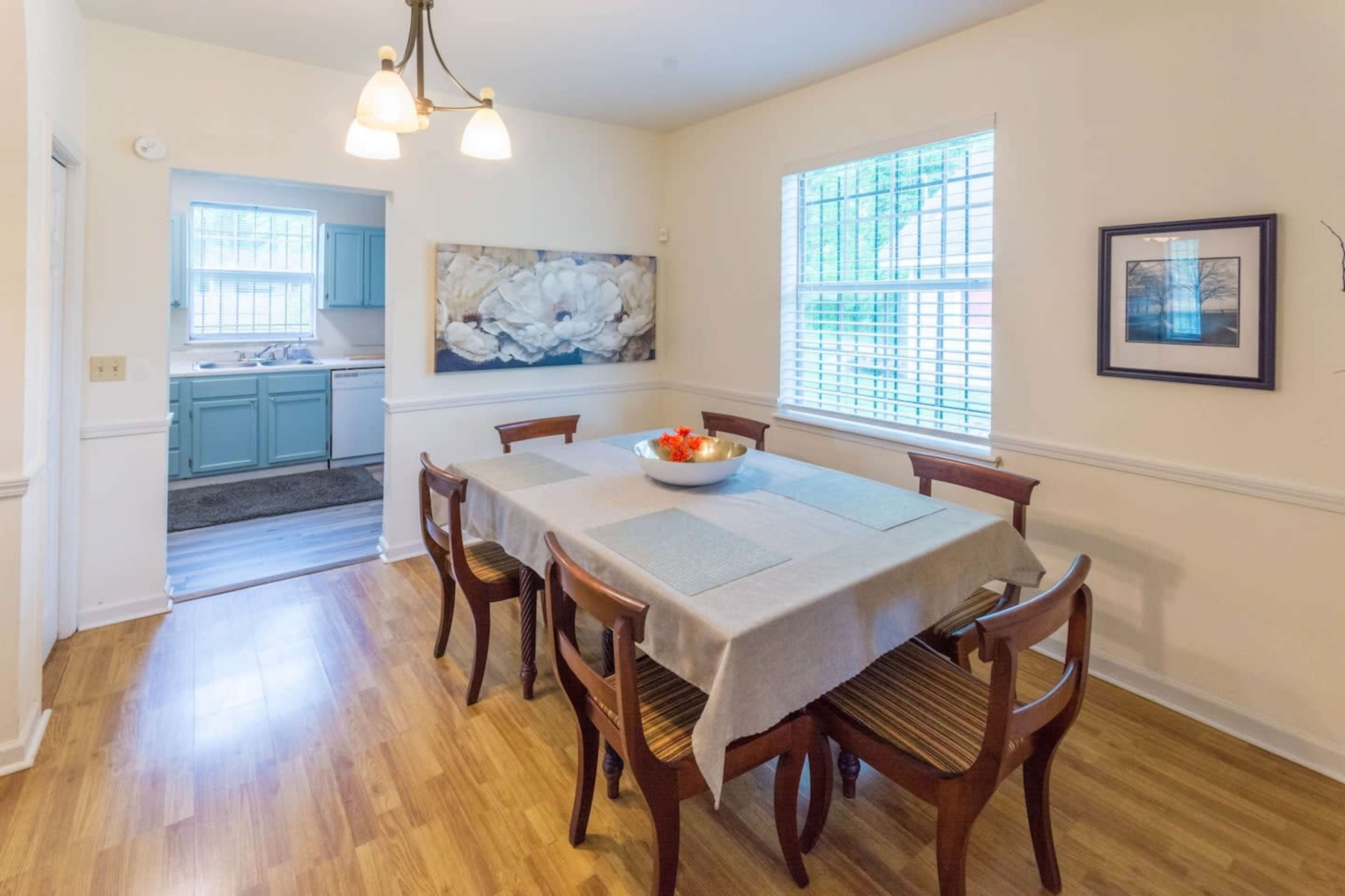 A dining area features a wooden table with eight chairs, a bowl of fruit in the center, and windows that provide natural light.