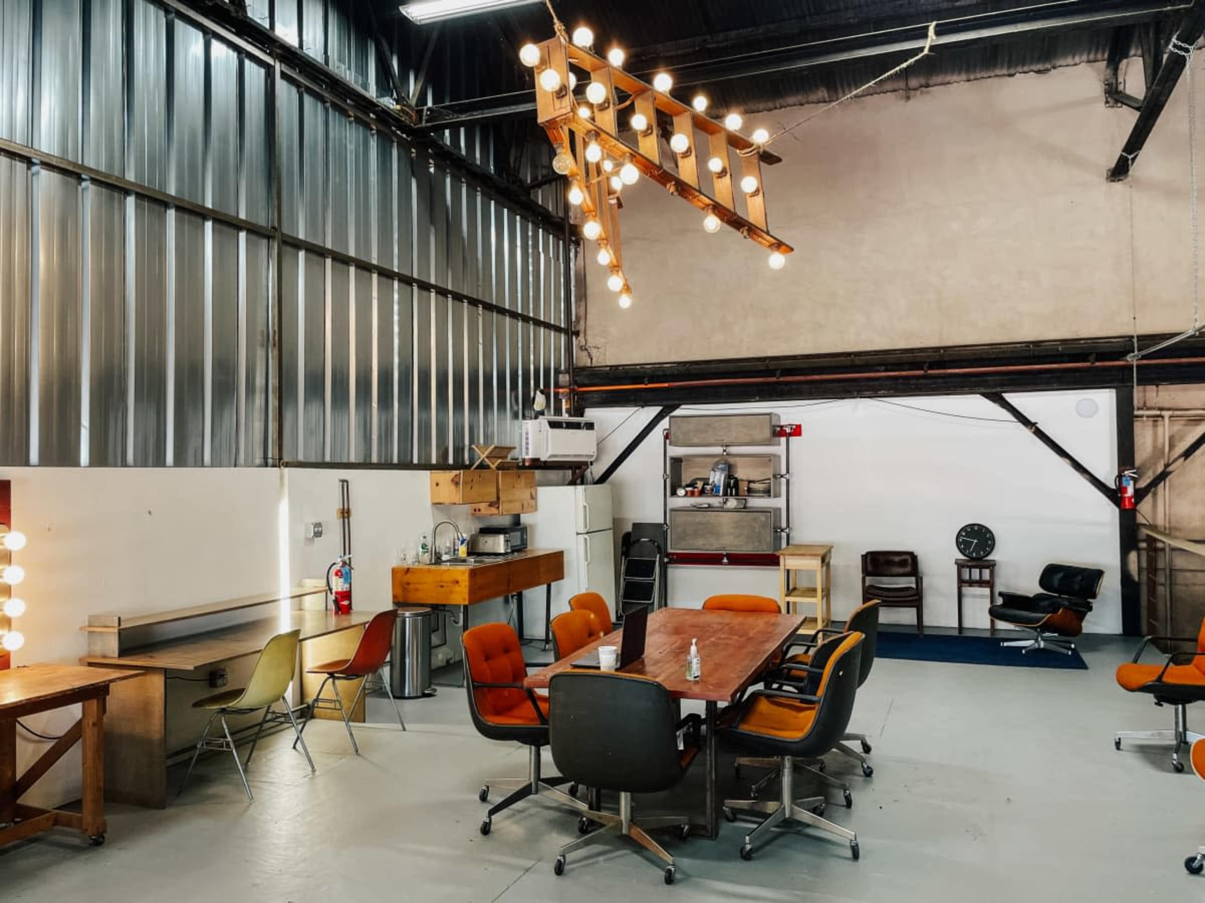 The image shows an industrial-style workspace featuring a long wooden table surrounded by colorful chairs, with a large light fixture hanging above.