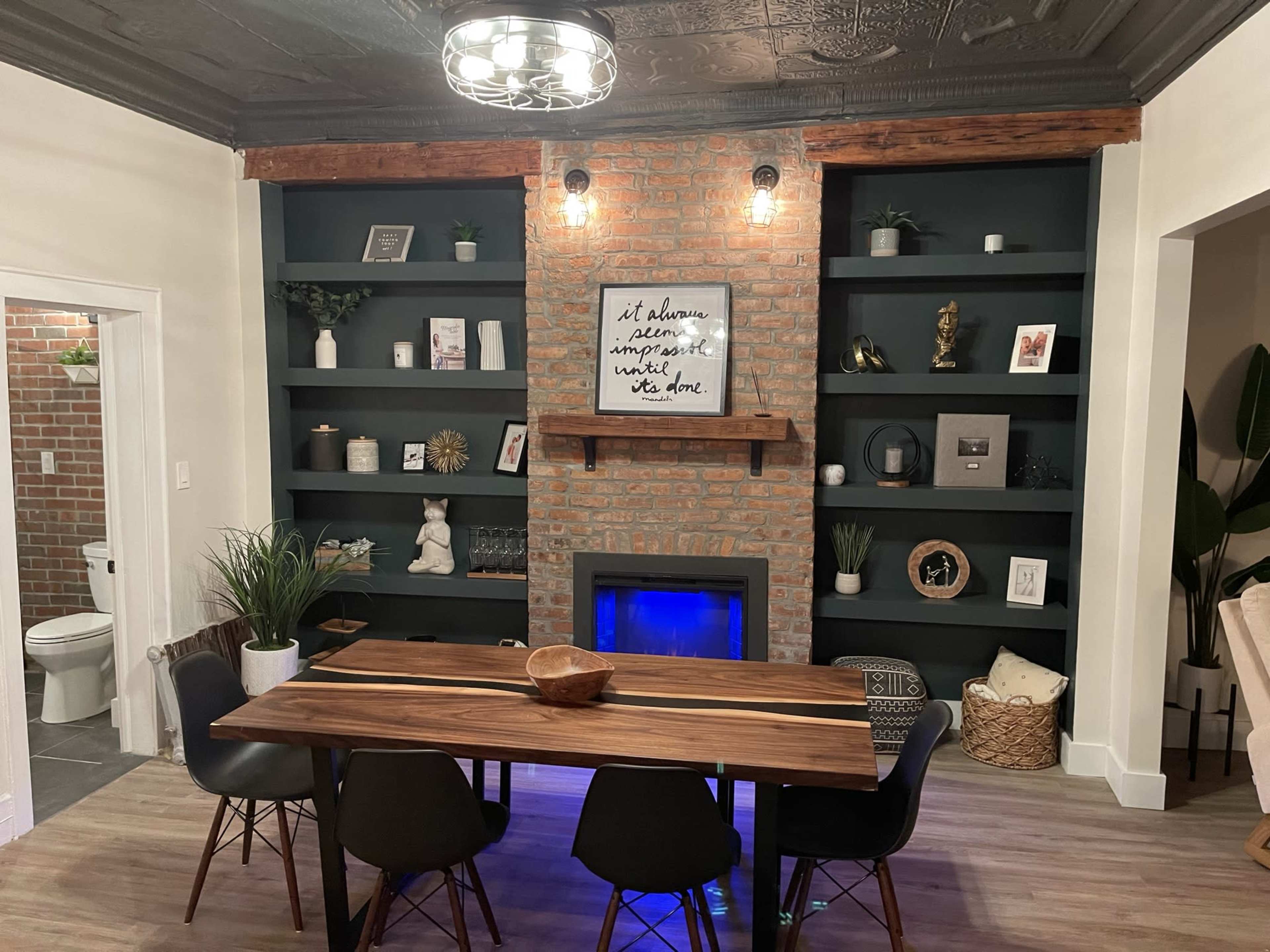 A dining area featuring a long wooden table, black chairs, a brick wall with shelves displaying decor, and an electric fireplace with blue lighting.
