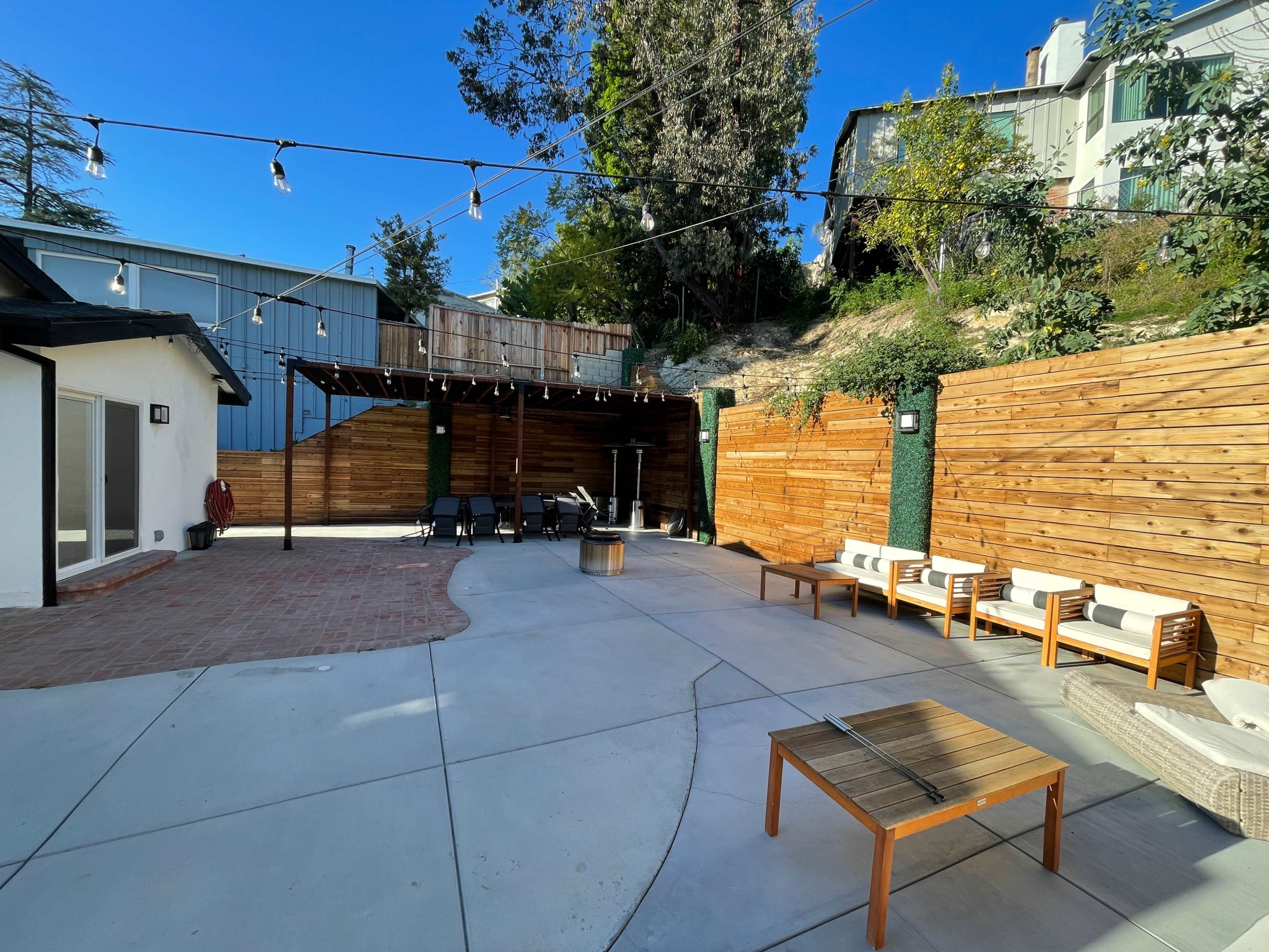 The image shows an outdoor patio area with concrete flooring, wooden walls, and a pergola, featuring seating arrangements and string lights above.