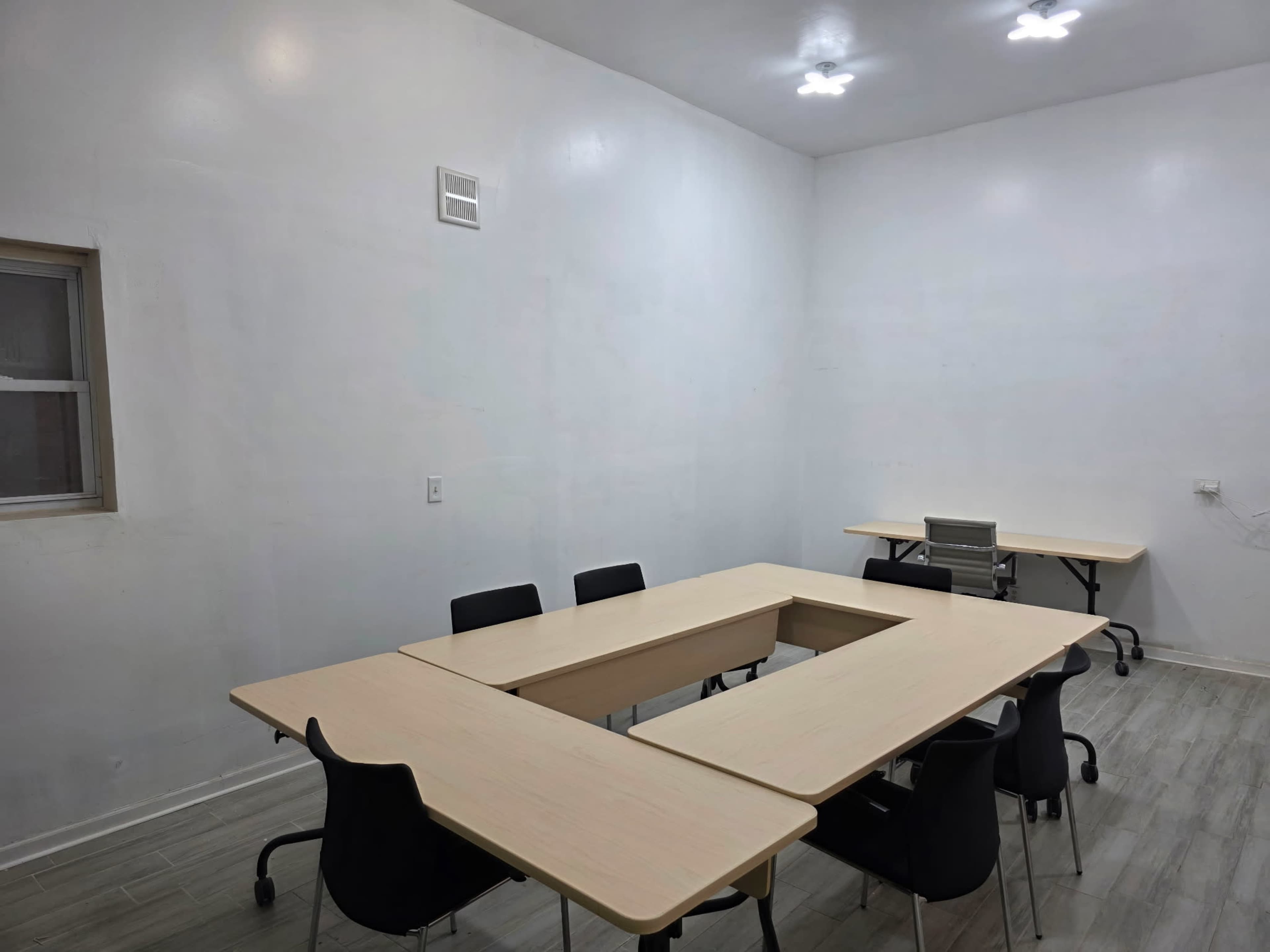 The image shows a sparsely furnished meeting room with a large table arranged in a U-shape, surrounded by black chairs, and a separate table against the wall.