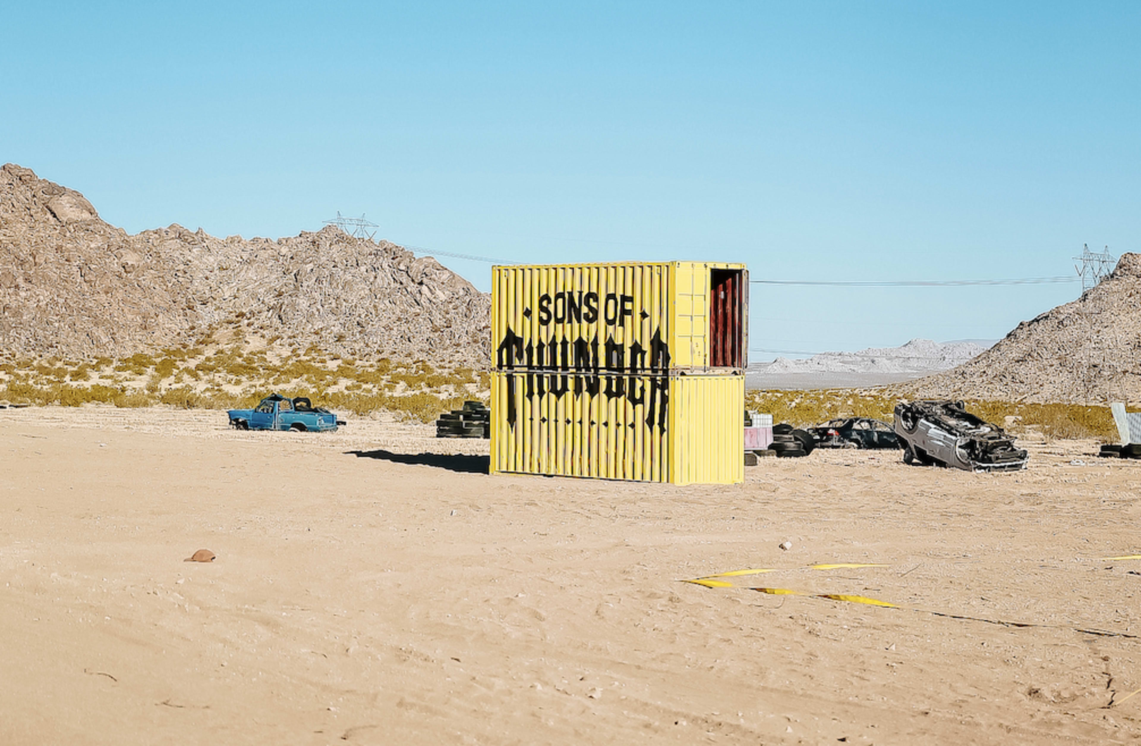 A yellow shipping container labeled "SONS OF" stands in a desert landscape surrounded by abandoned cars and rocky terrain.