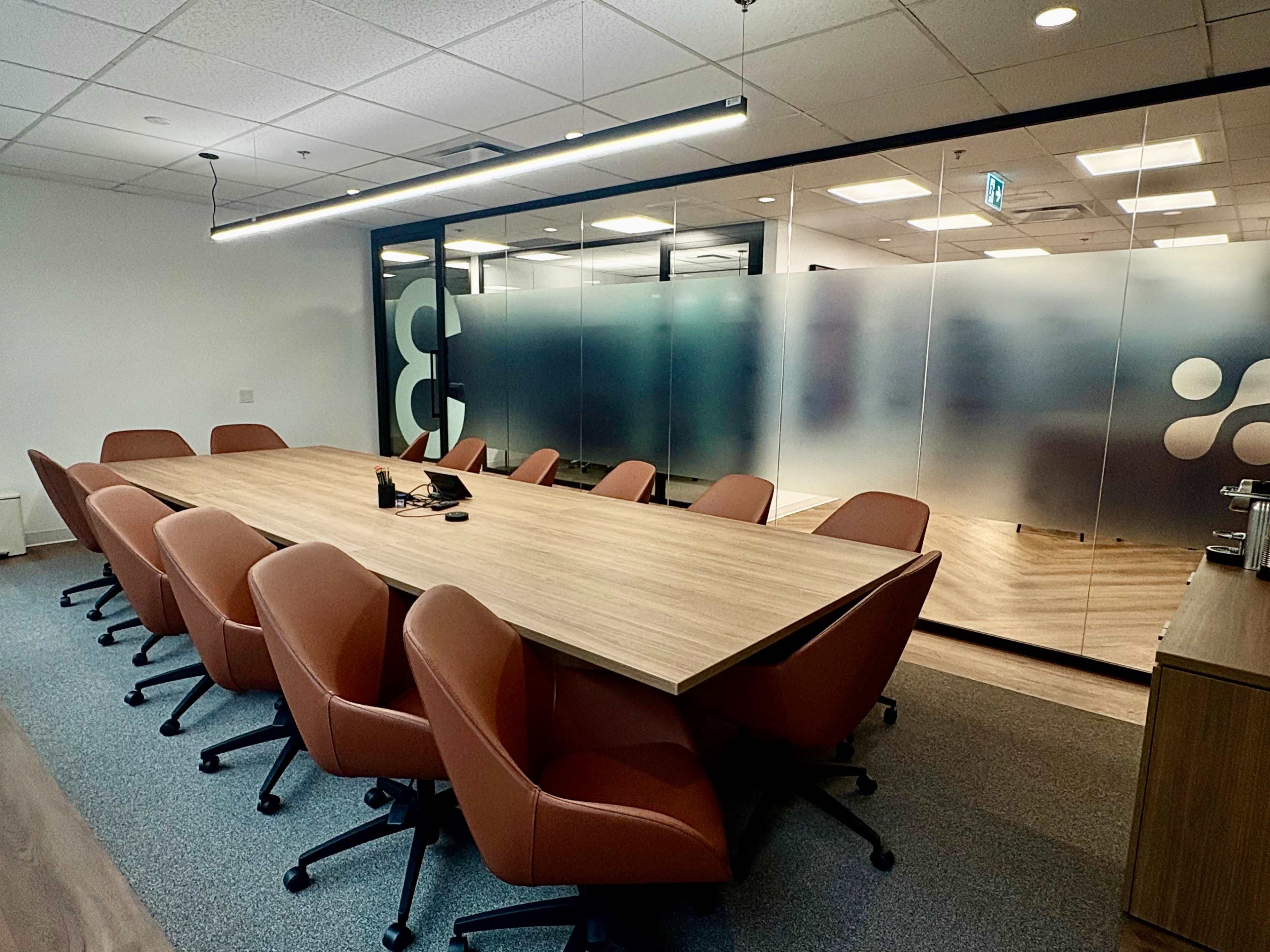 A conference room features a long wooden table surrounded by brown office chairs, with a glass wall partitioning a separate meeting area.