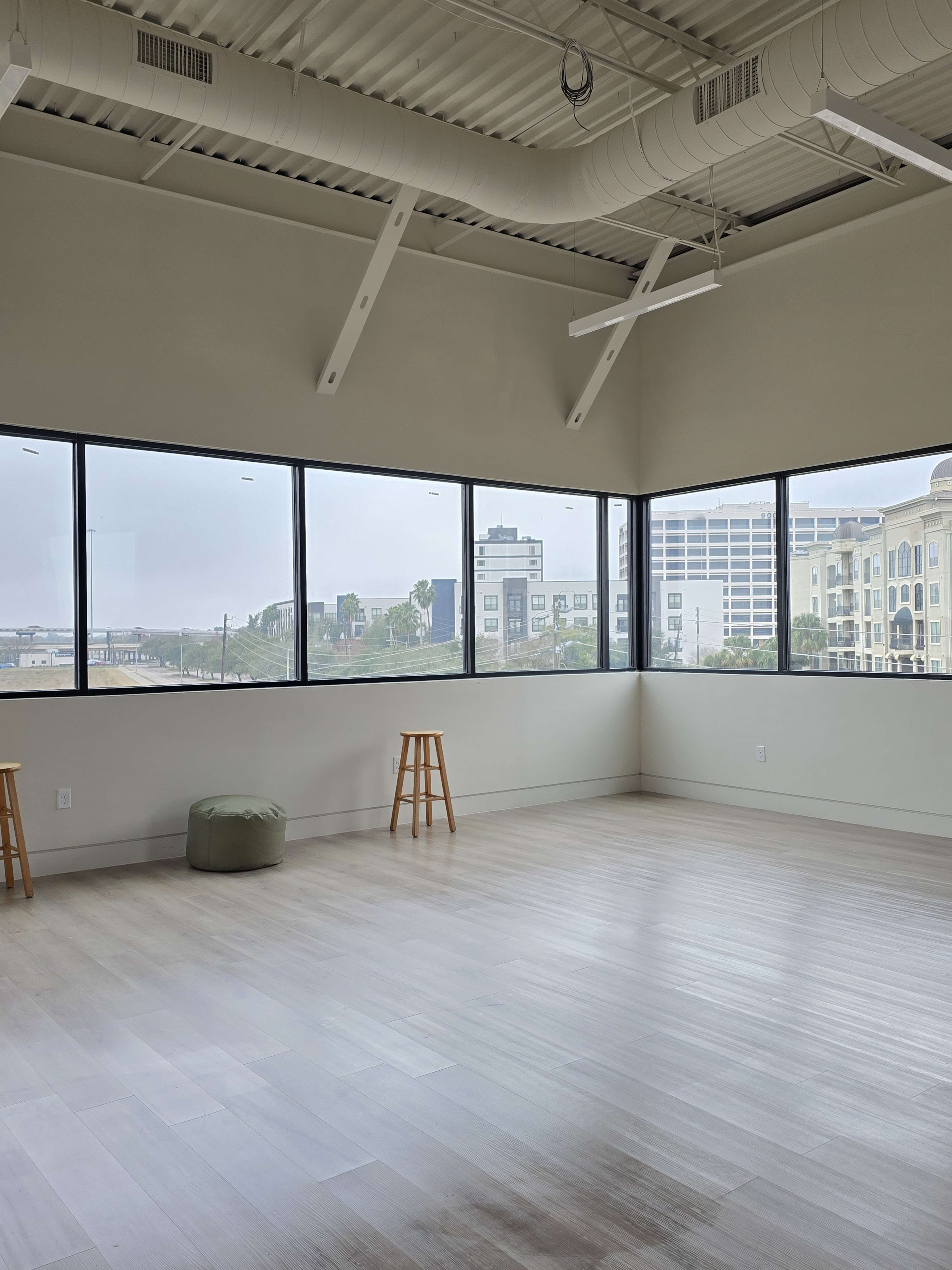 The image shows a spacious room with wooden floors, large windows, and two wooden stools placed near the windows, overlooking an urban landscape.