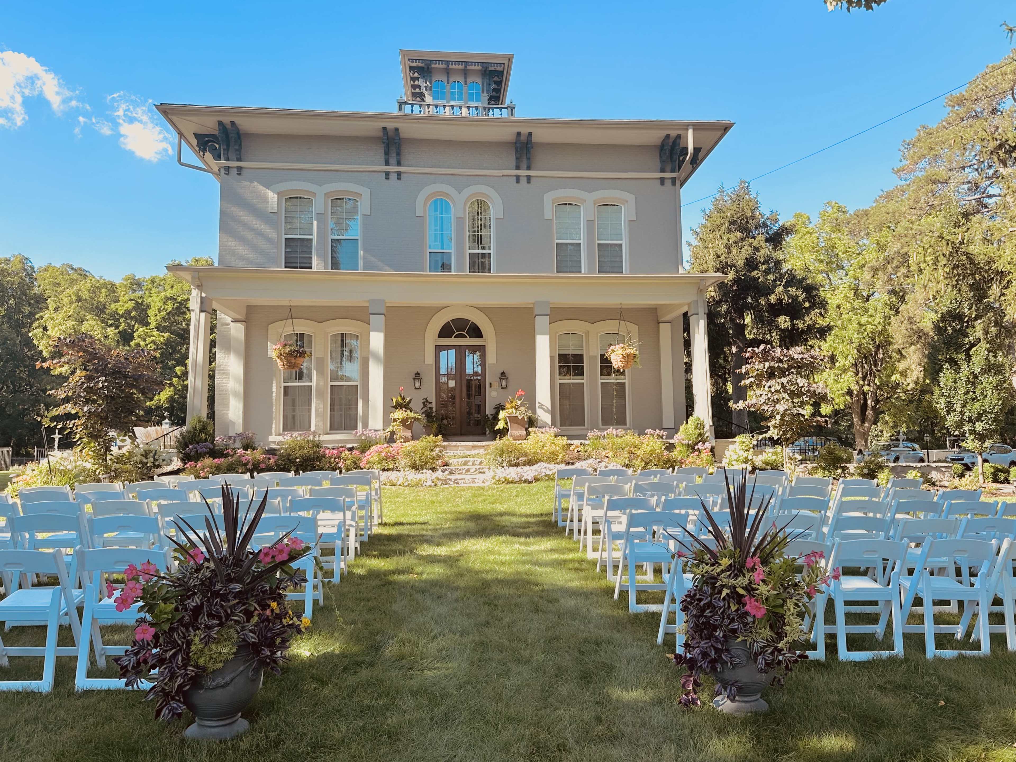 A large, elegant house with a front porch, surrounded by neatly arranged chairs set up for an outdoor event in a landscaped yard.