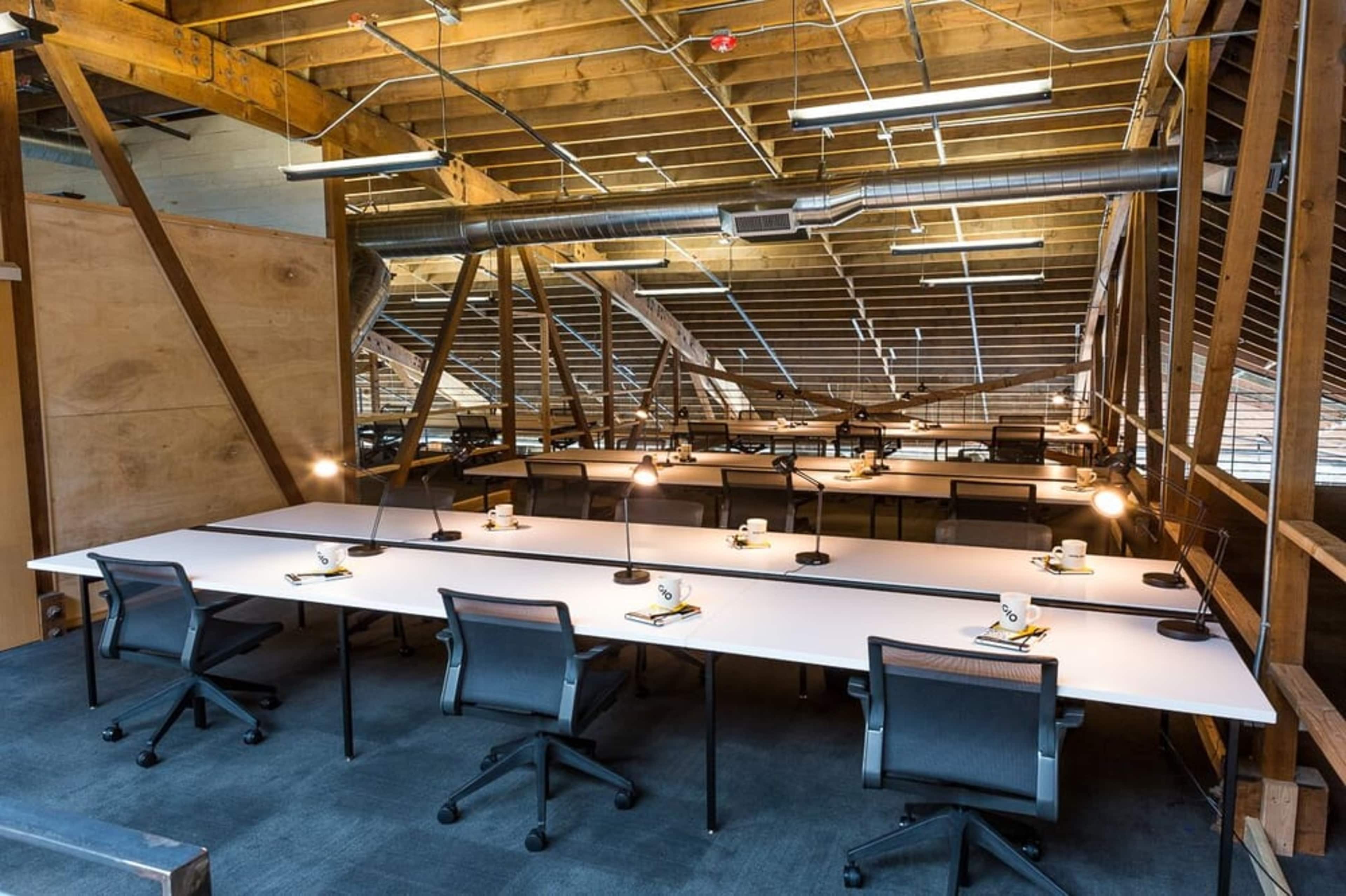 A spacious room with multiple workstations lined with white desks, each equipped with lamps and chairs, under a wooden ceiling structure.