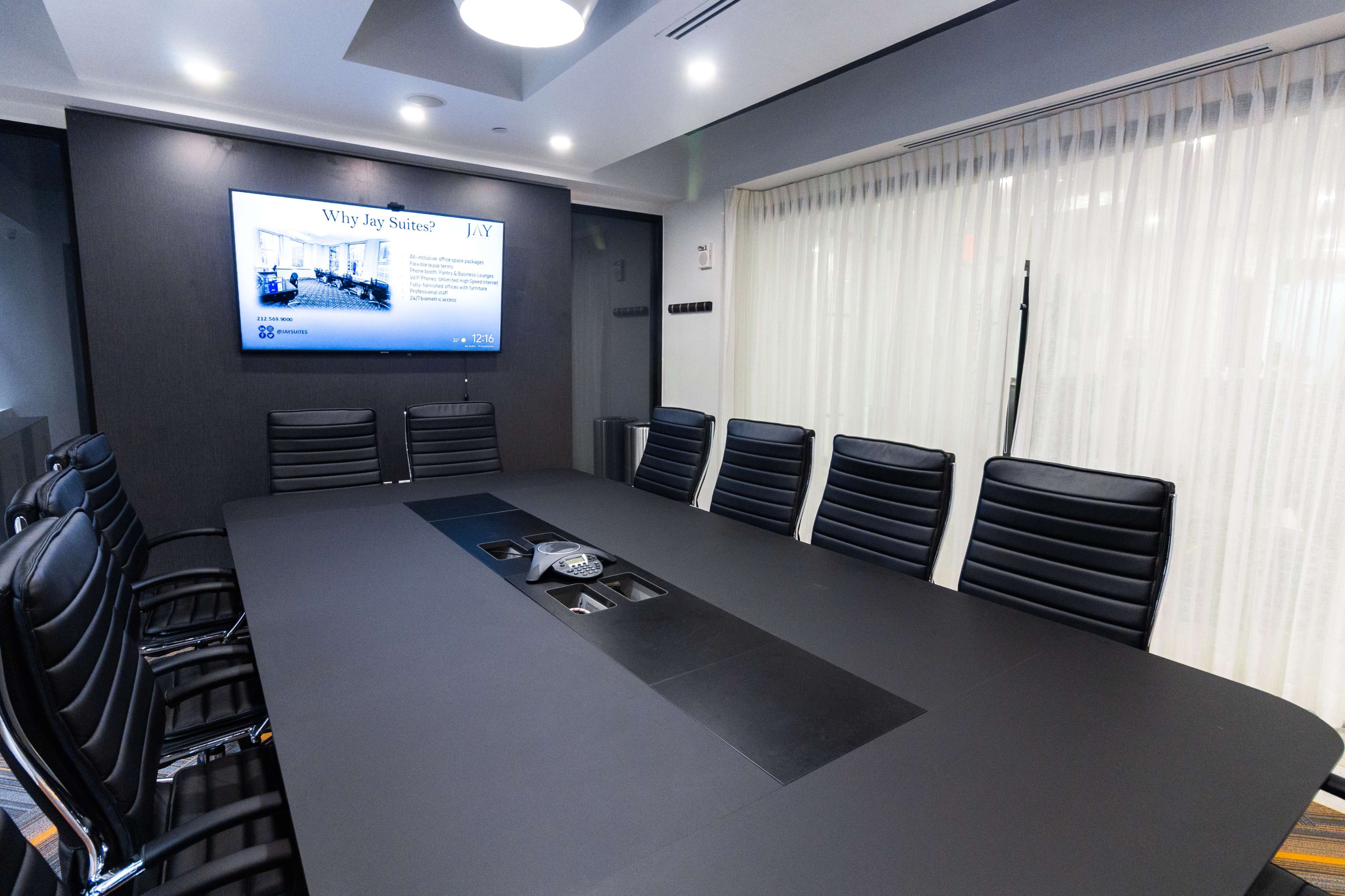 A large conference room features a long table surrounded by black leather chairs, with a screen displaying a presentation on the wall.