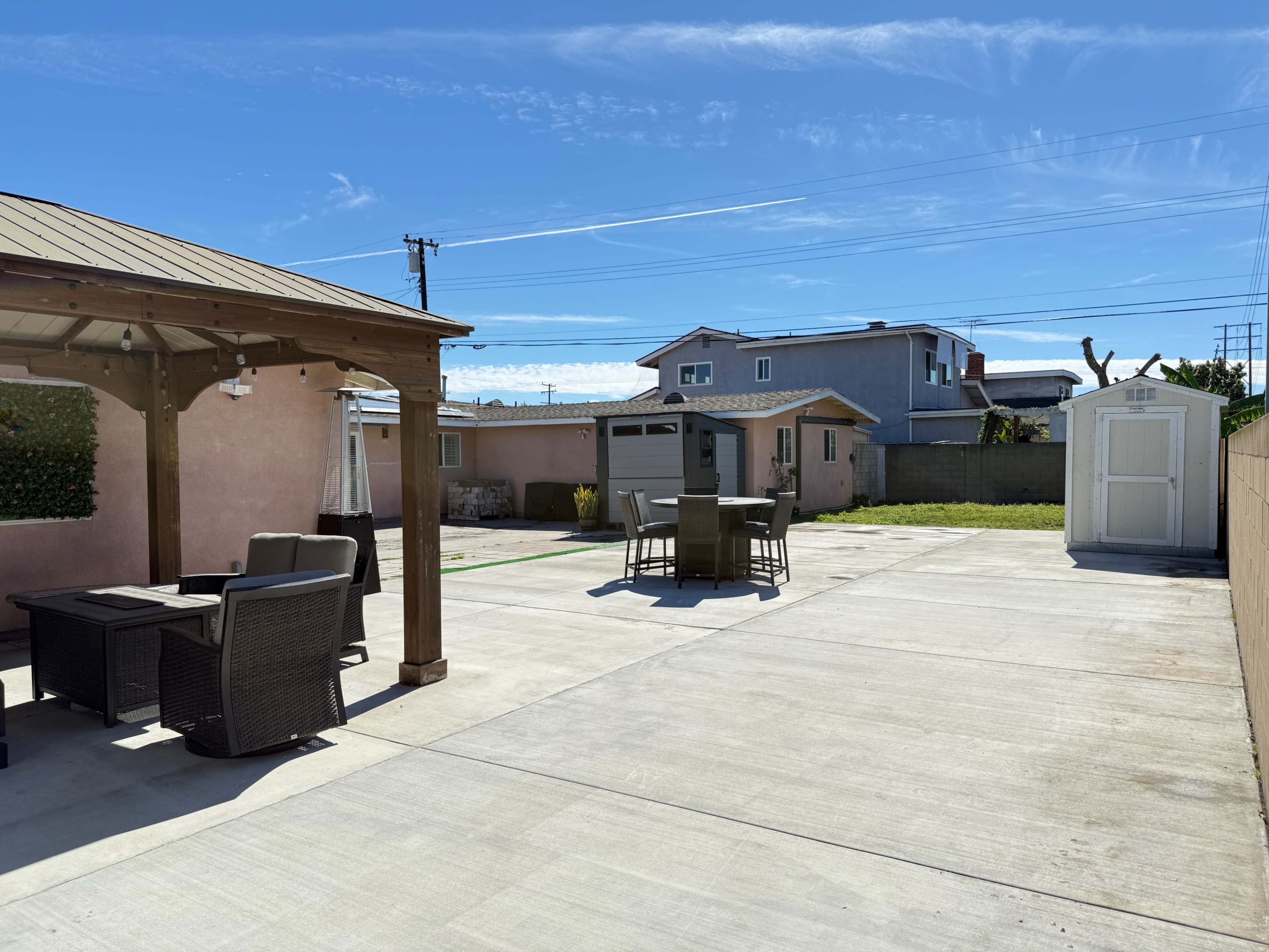 The image shows a flat, paved backyard with a wooden gazebo, outdoor seating, a dining area, and a storage shed in clear weather.