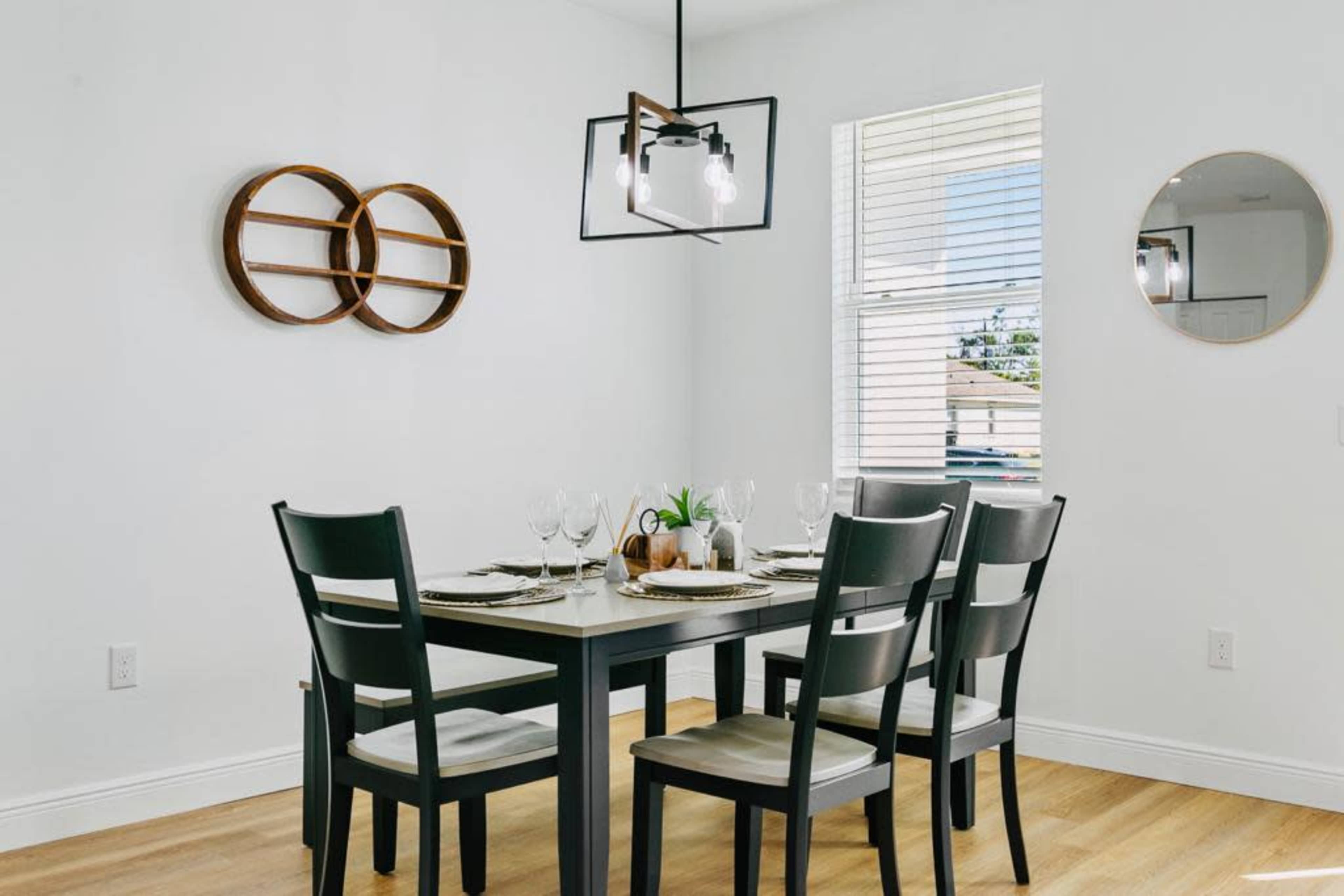 A modern dining area features a table set for four with black chairs, a minimalist light fixture, and wall decor including shelves and a round mirror.