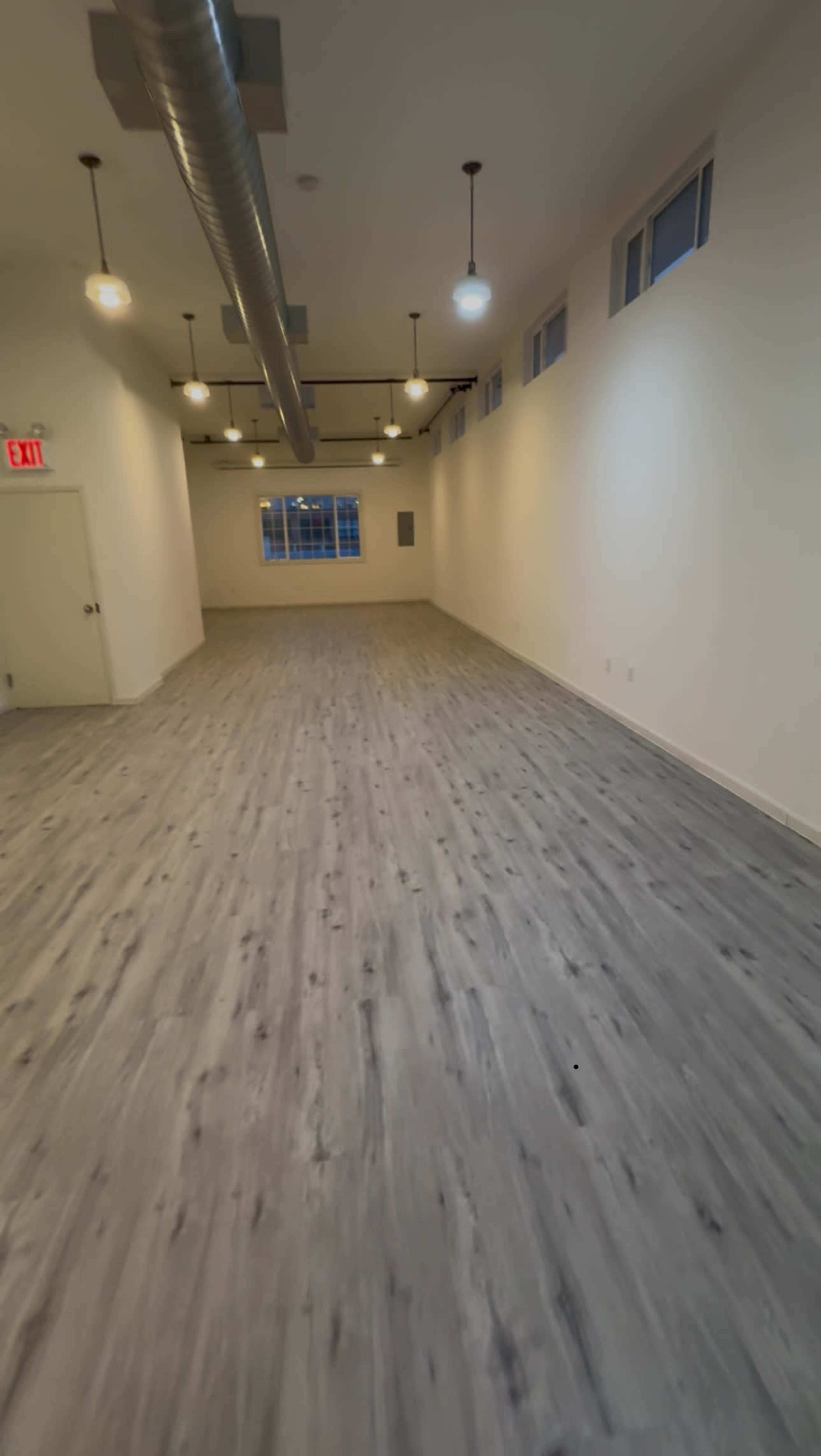 The image shows a spacious, empty room with light-colored walls, wooden flooring, and overhead lighting fixtures.