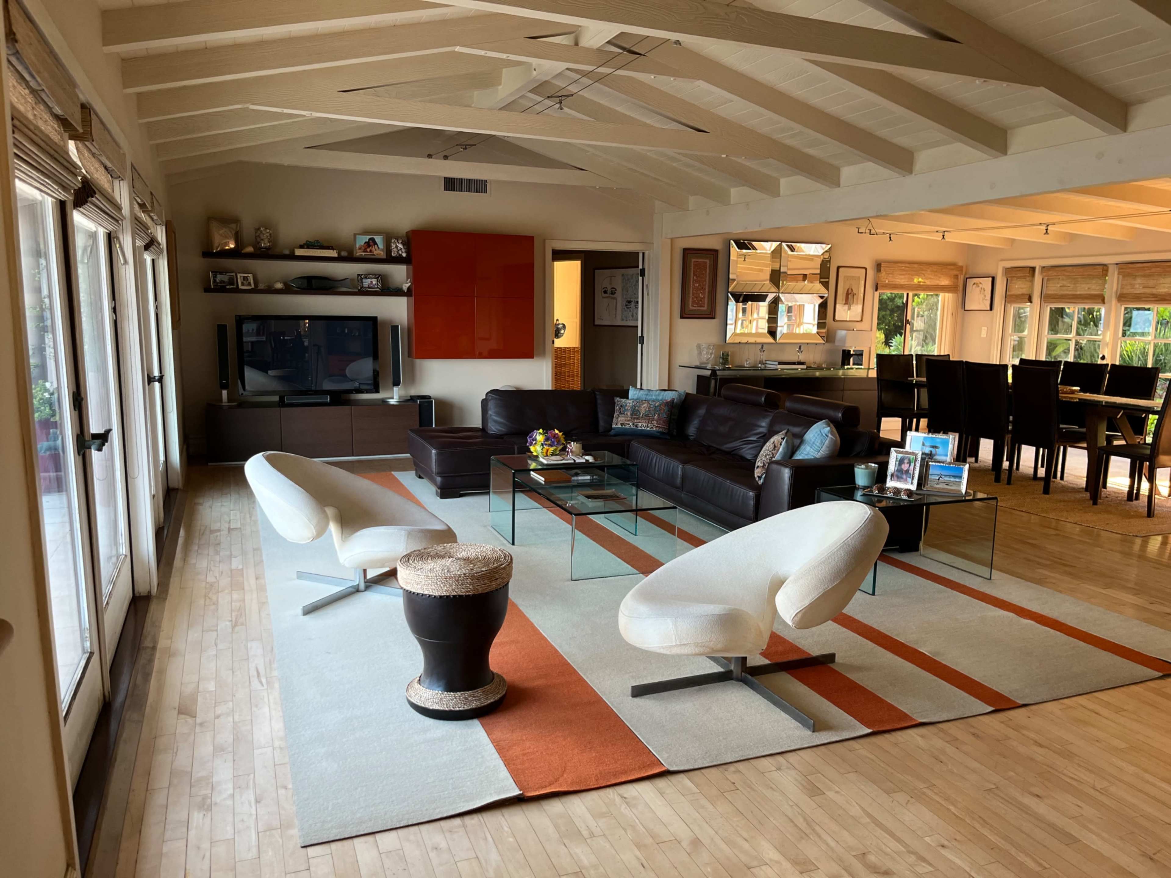 The image shows a spacious living area with modern furniture, including a brown leather sofa and two white chairs, separated by a glass coffee table on a large area rug.