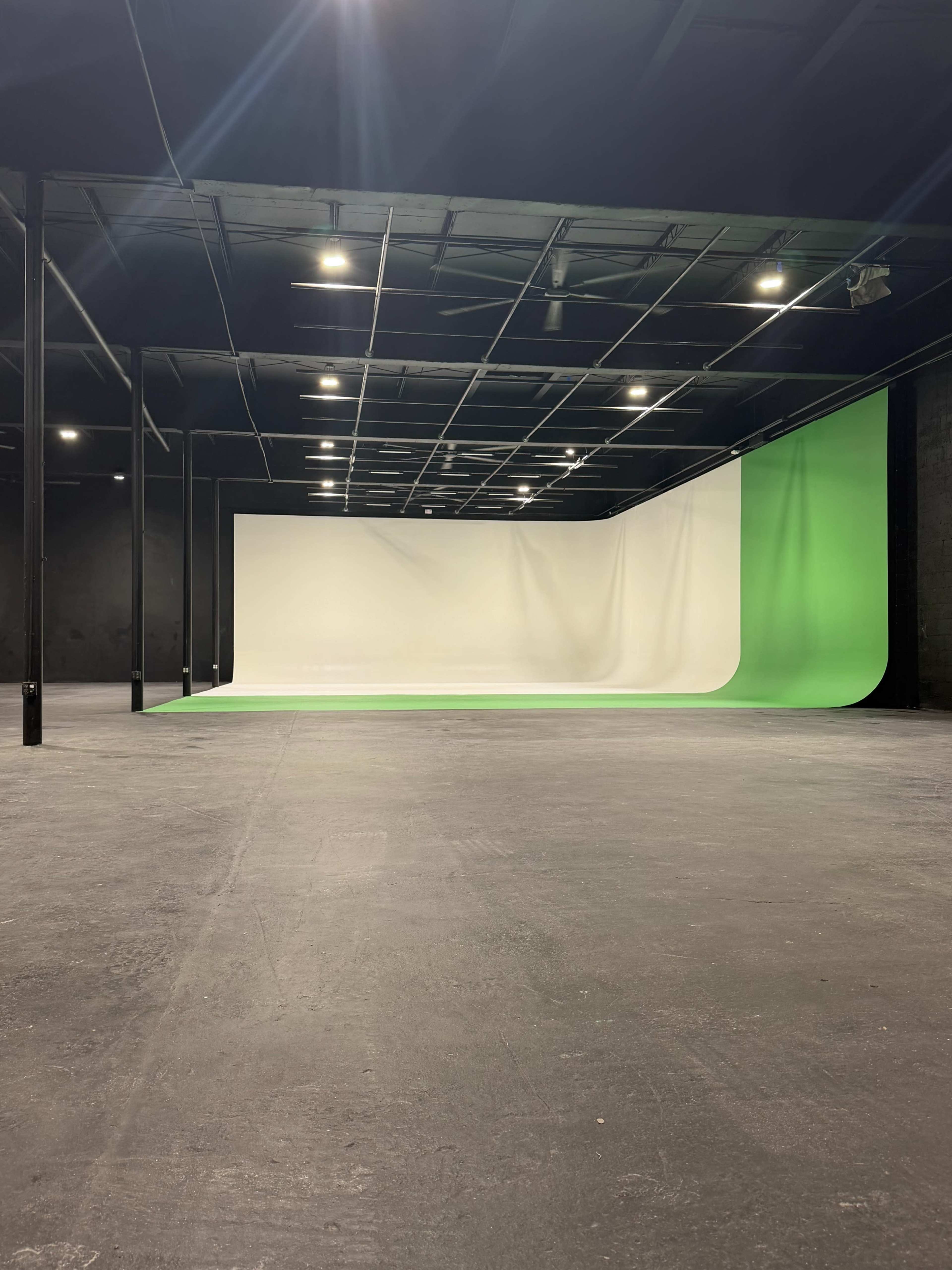 The image shows a spacious studio with a black ceiling, concrete floor, and a large seamless backdrop that transitions from white to green.