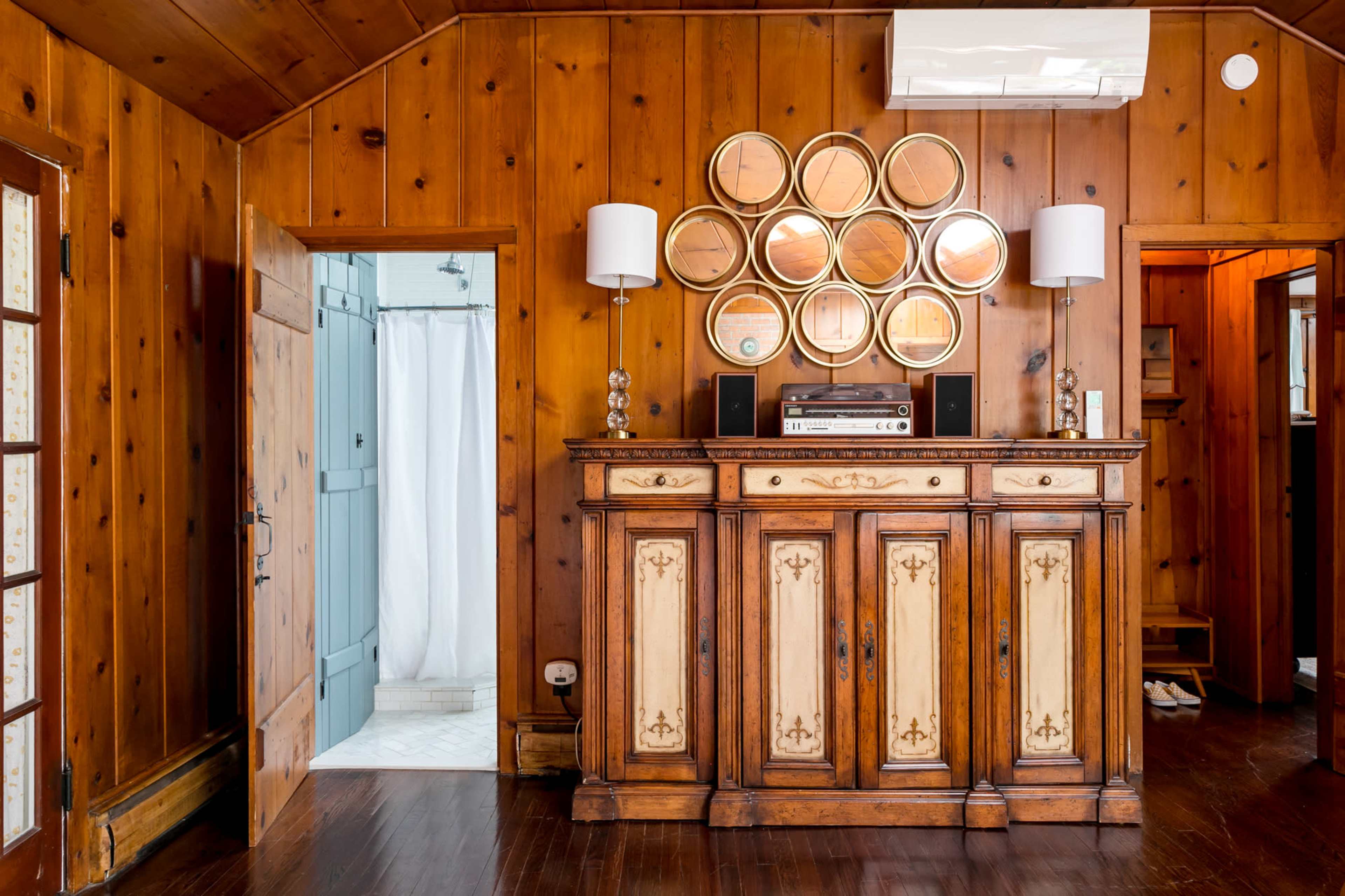 The image shows a wooden-paneled room with a vintage cabinet adorned with circular mirrors and two lamps, while a doorway leads to a bathroom with a shower curtain visible.