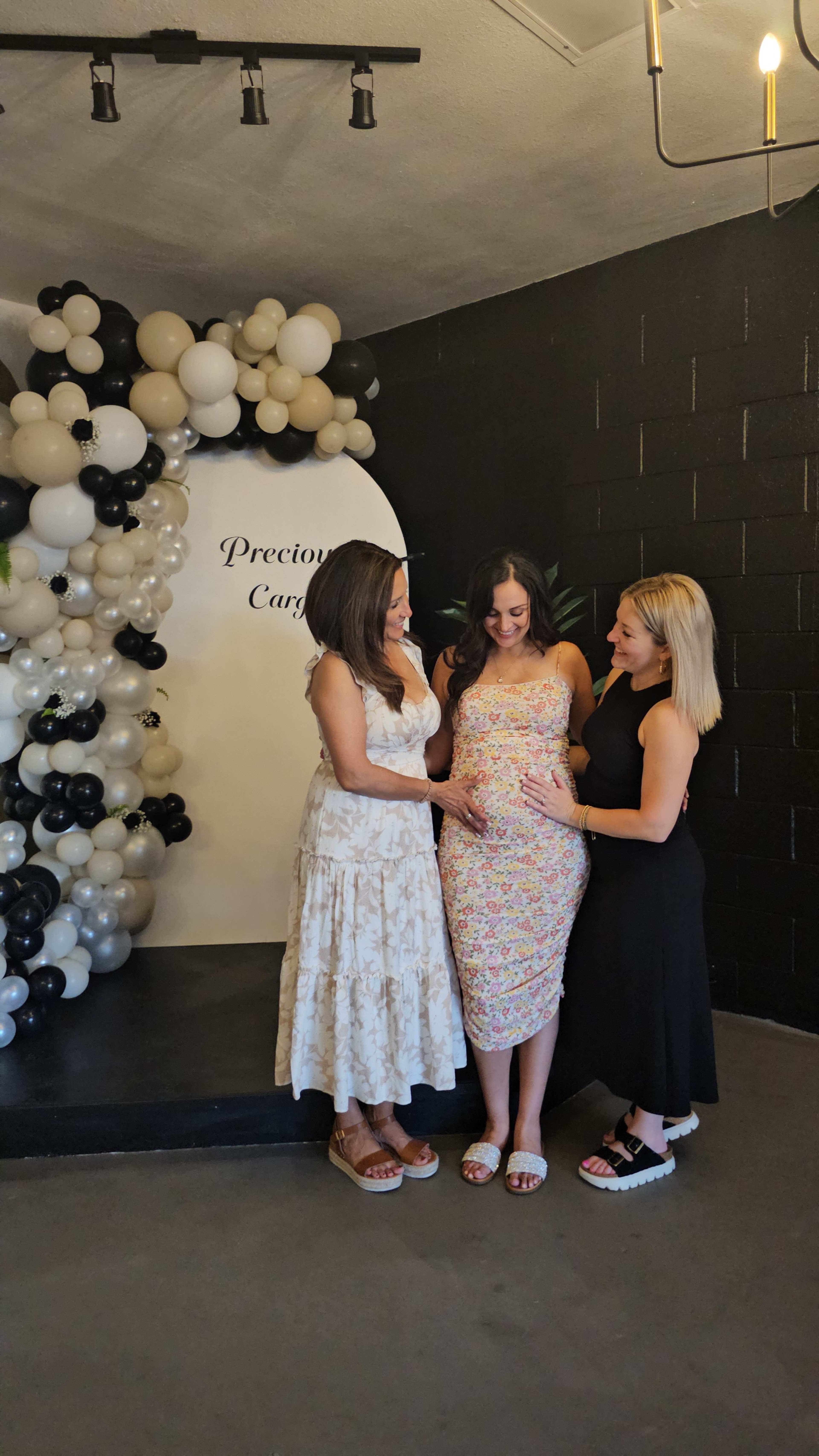 Three women stand together in front of a balloon arch, with one woman visibly pregnant.
